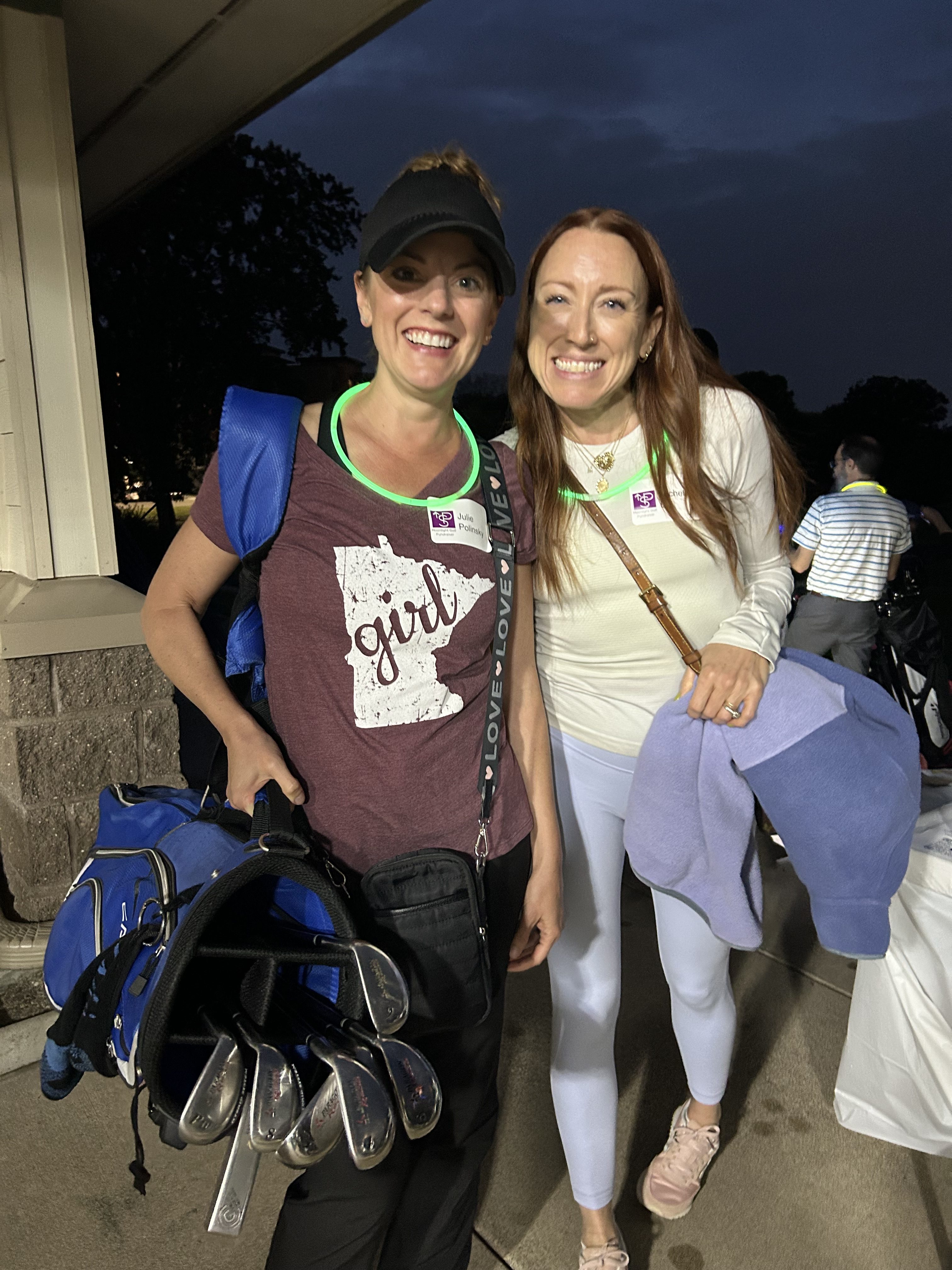 Two women standing together outdoors at night, one holding golf clubs and smiling, both wearing glow necklaces.