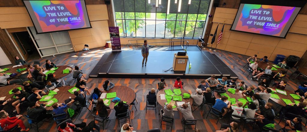 A speaker stands on stage addressing an audience in a conference room with two screens displaying a motivational message.