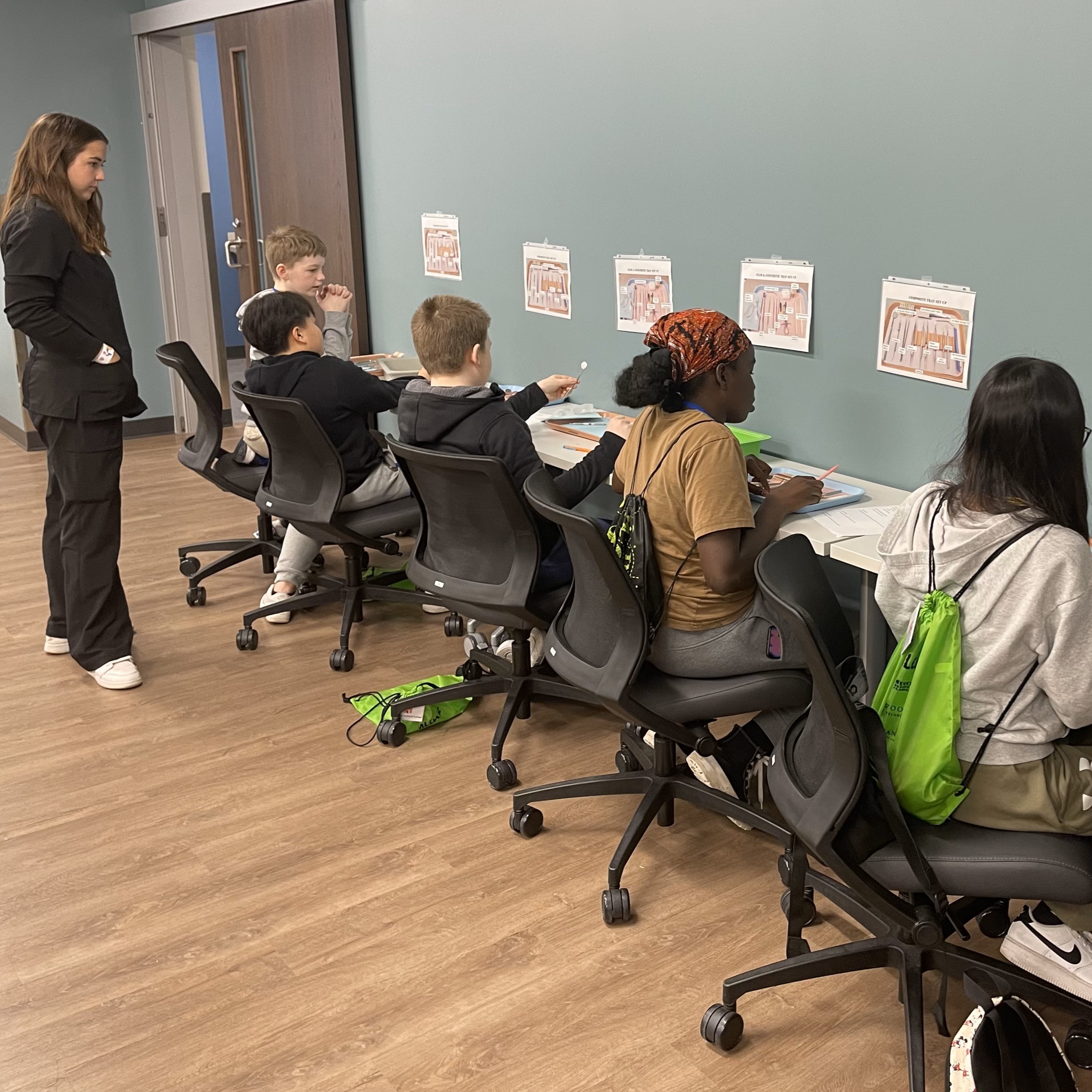 Several students sit a long table working with papers while an professor observes in a classroom with green walls and wood flooring.