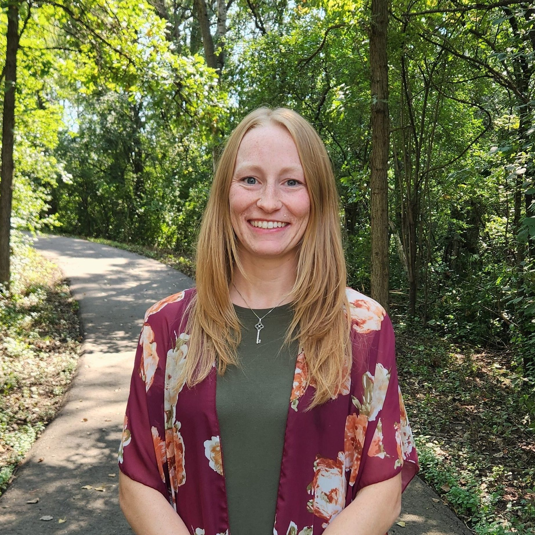 A woman with long hair stands smiling on a forested path, wearing a floral kimono over a green top.