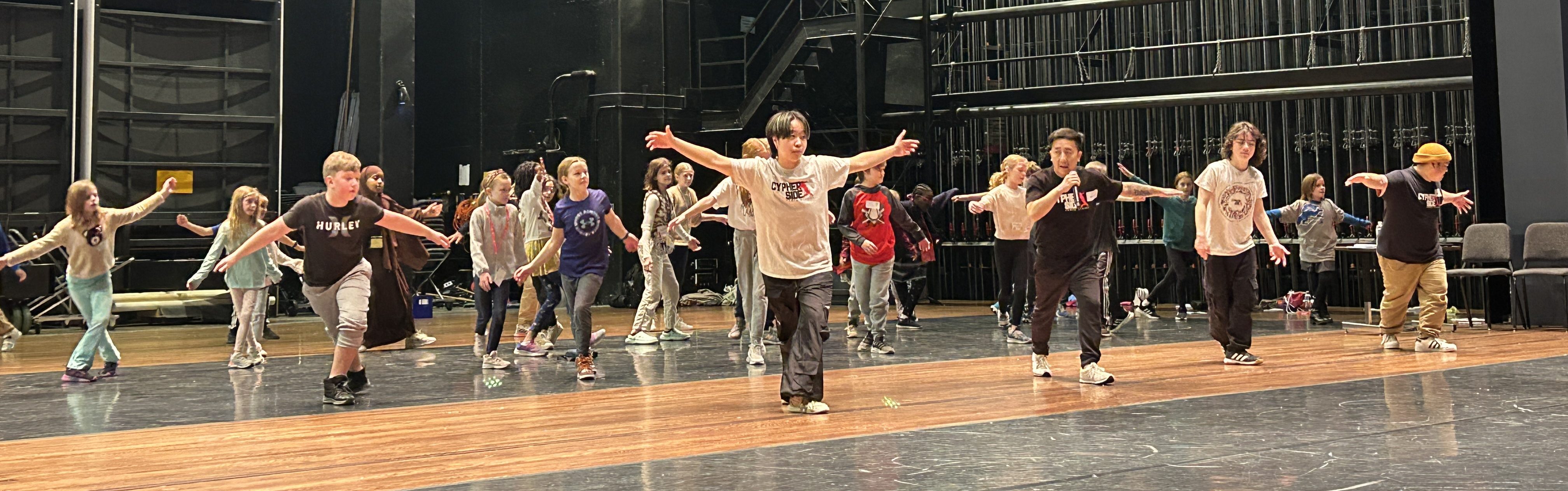 Children practicing a breakdance routine in a large theater rehearsal space with black walls and bright lights.