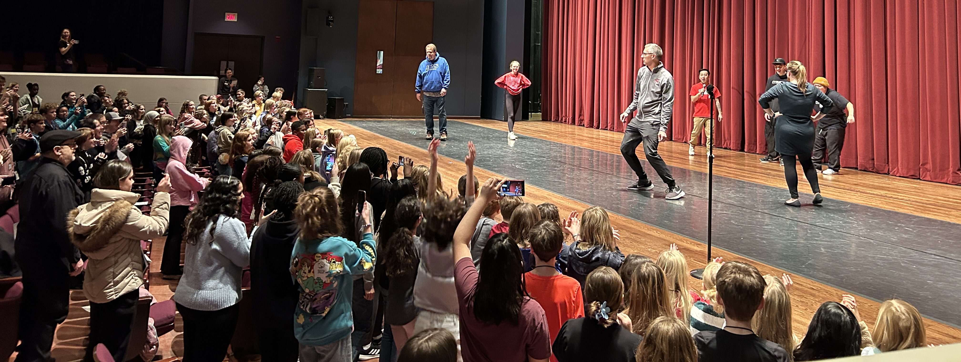Teachers and students having a dance-off on stage with red curtains, audience watching and cheering in a theater setting.