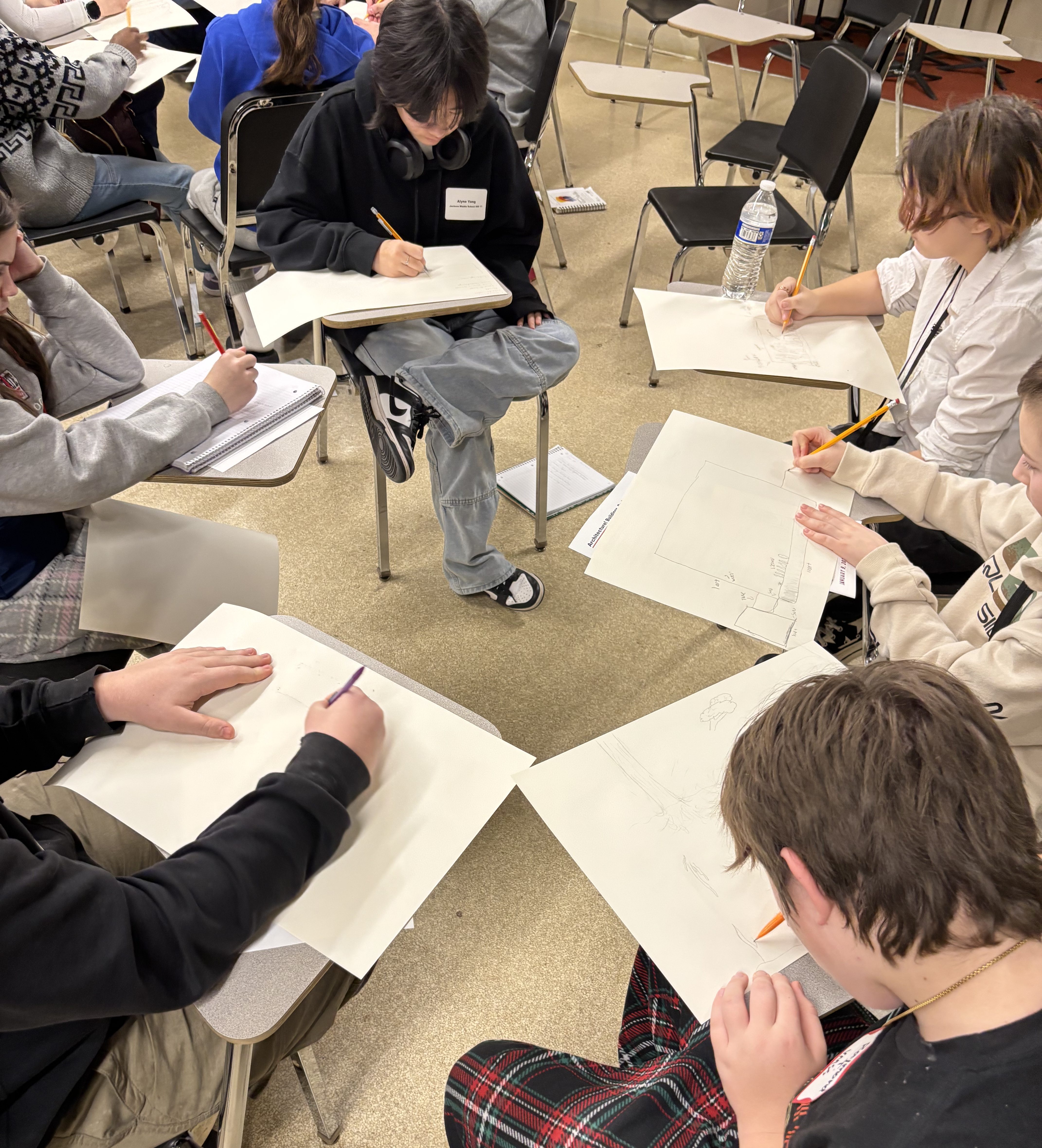 A group of students seated in a circle, sketching on large white paper with pencils in a classroom setting.