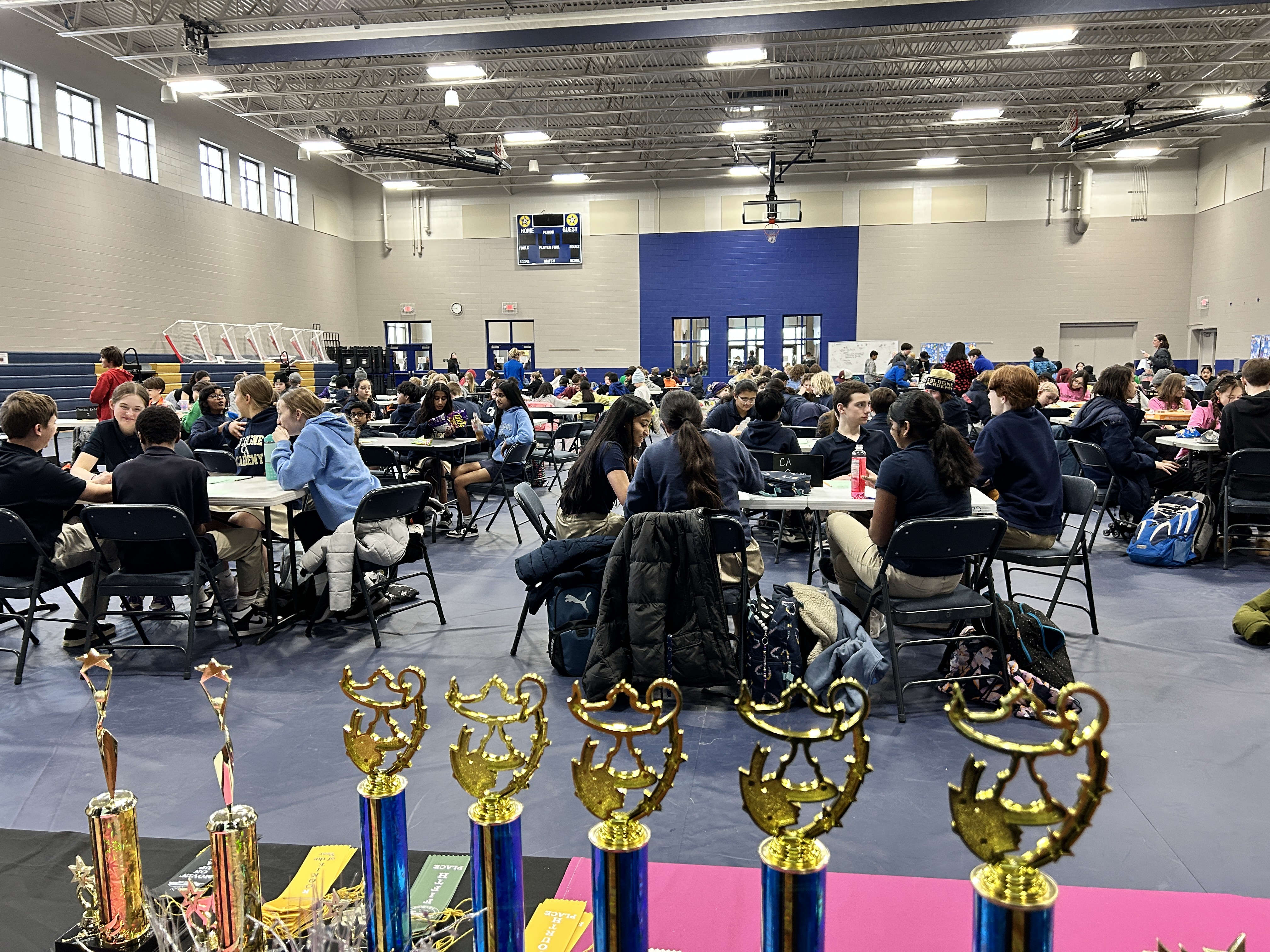 Knowledge Bowl teams at tables in a large hall with trophies displayed in the foreground.