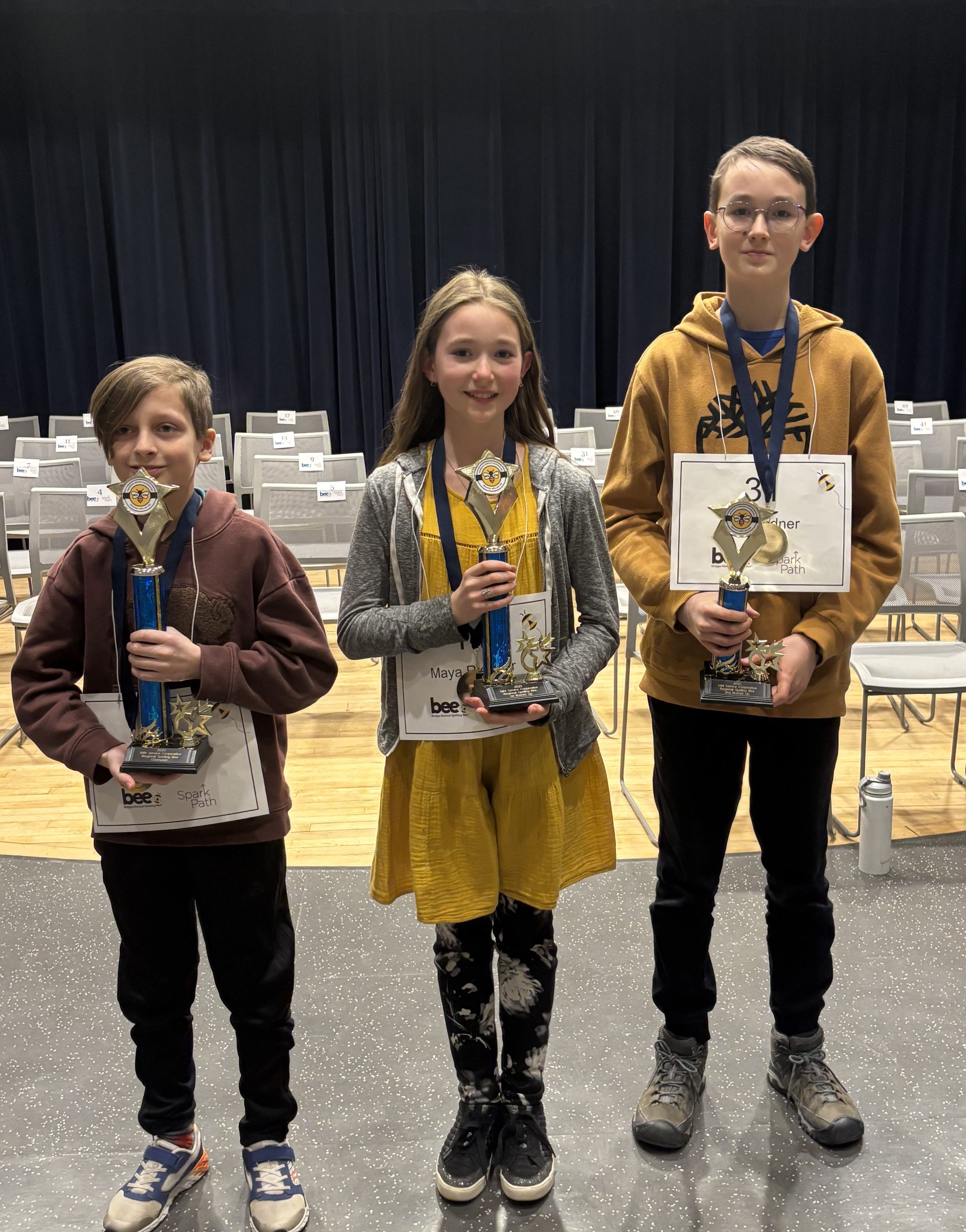 Three children standing on a stage holding trophies, with empty chairs in the background.