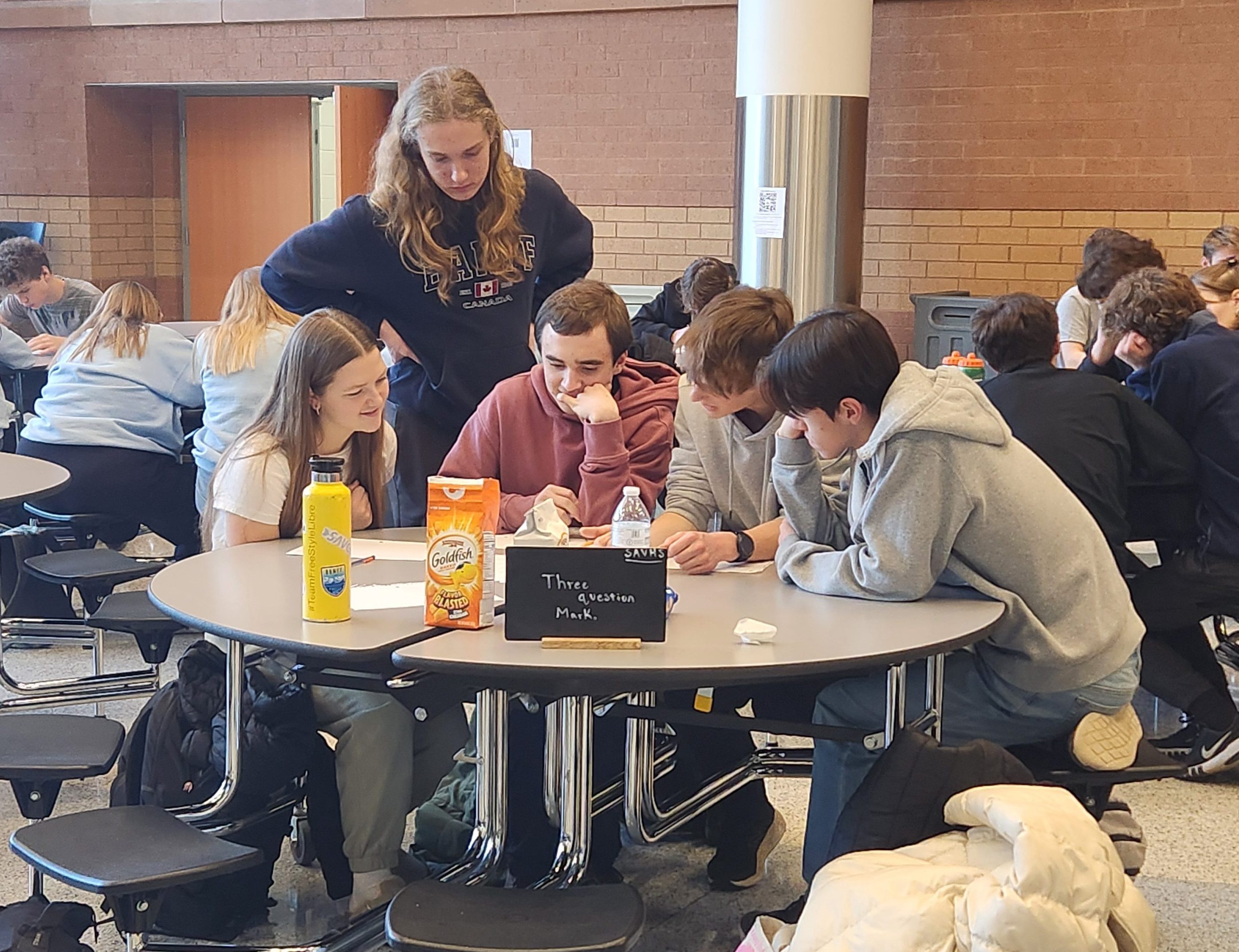 Four students sit around a table with snacks, collaborating on a quiz in a busy cafeteria.