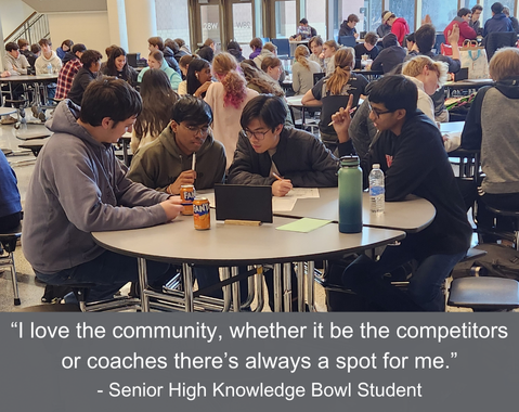 Four students work together at a table in a busy school cafeteria during a Knowledge Bowl competition.