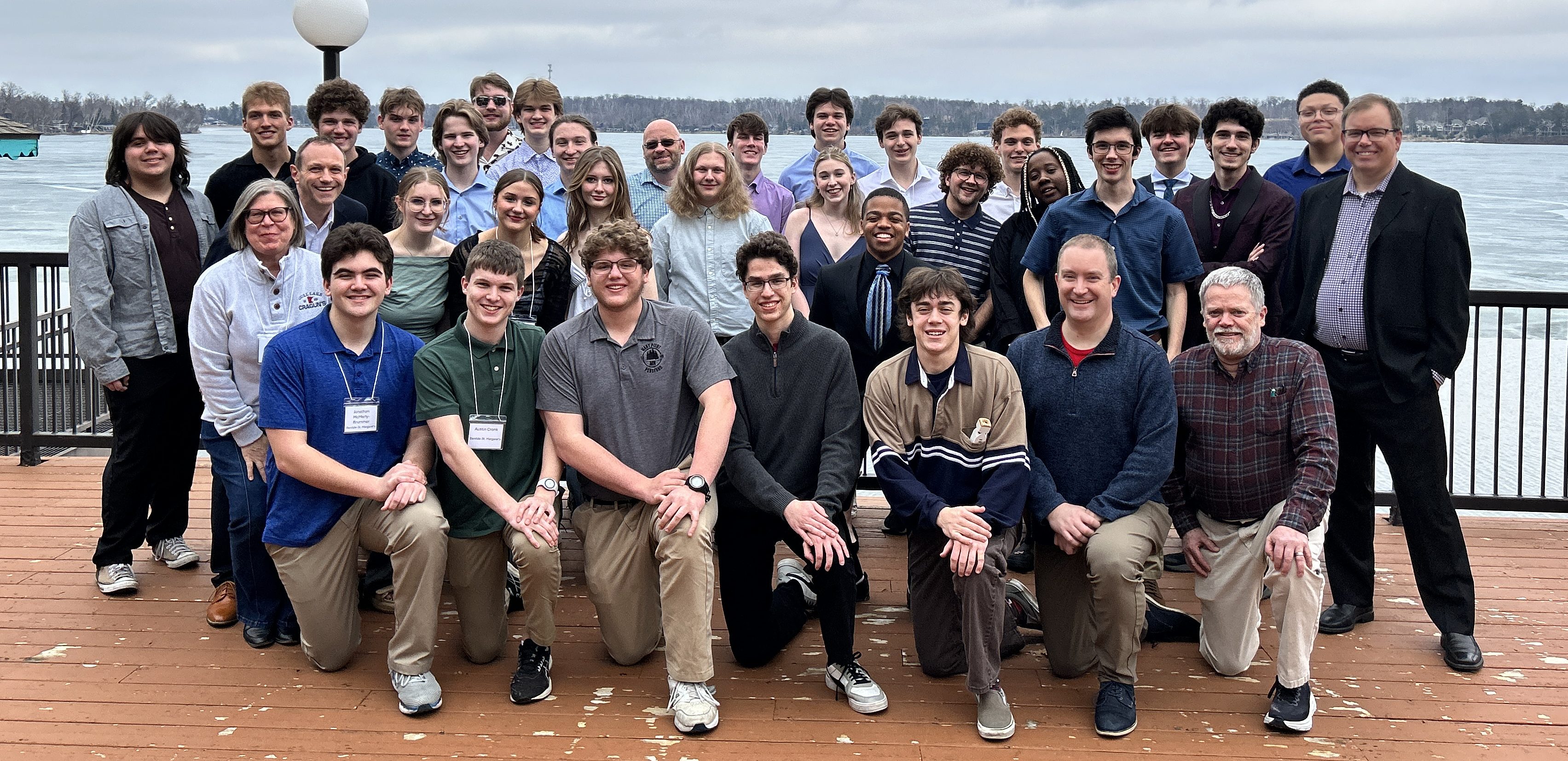 A group of people posing and smiling on a lakeside deck with cloudy skies and water in the background.