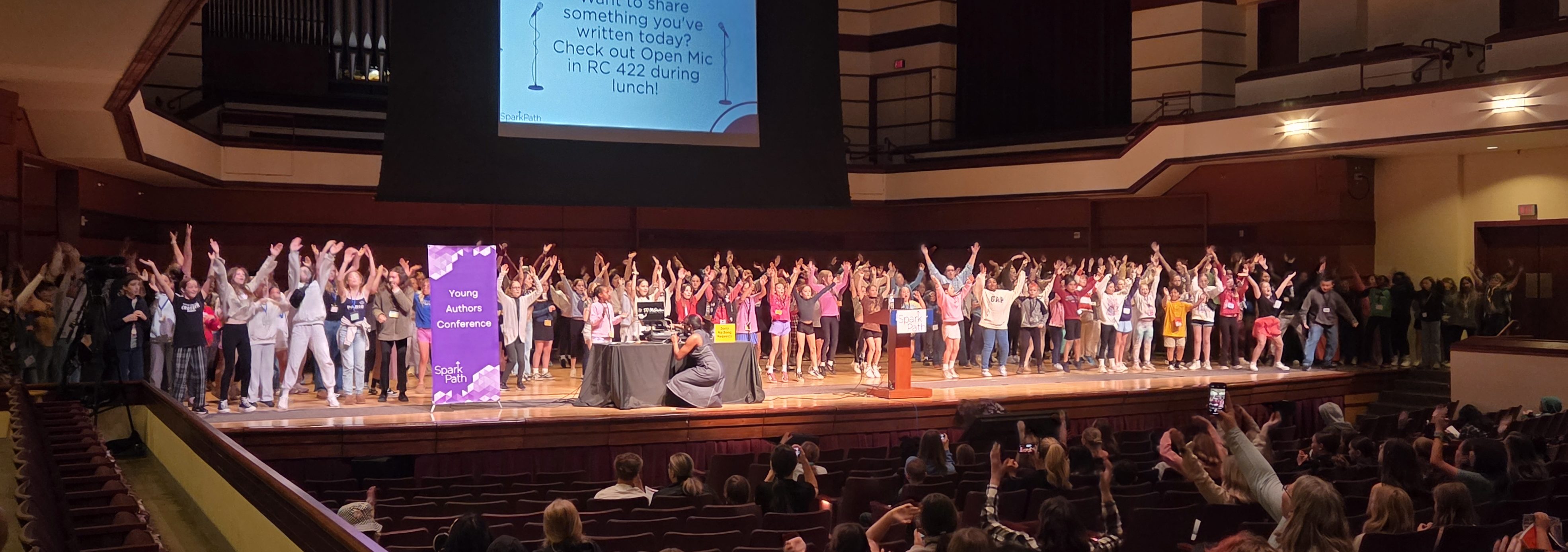 A large group of people stand on stage with arms raised, facing an audience in an auditorium.