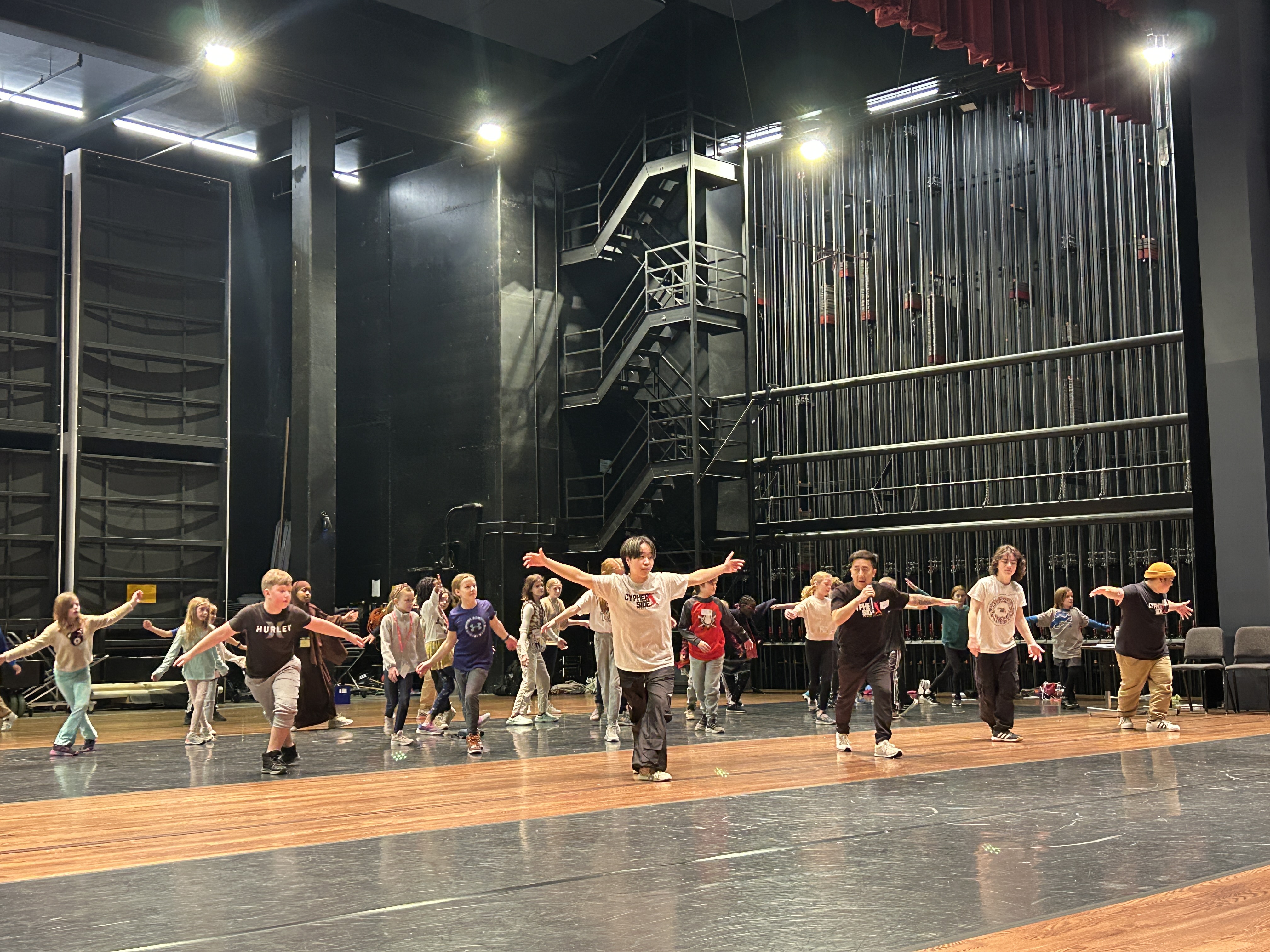 Children practicing a dance routine with arms outstretched on a large stage in a theater rehearsal space.