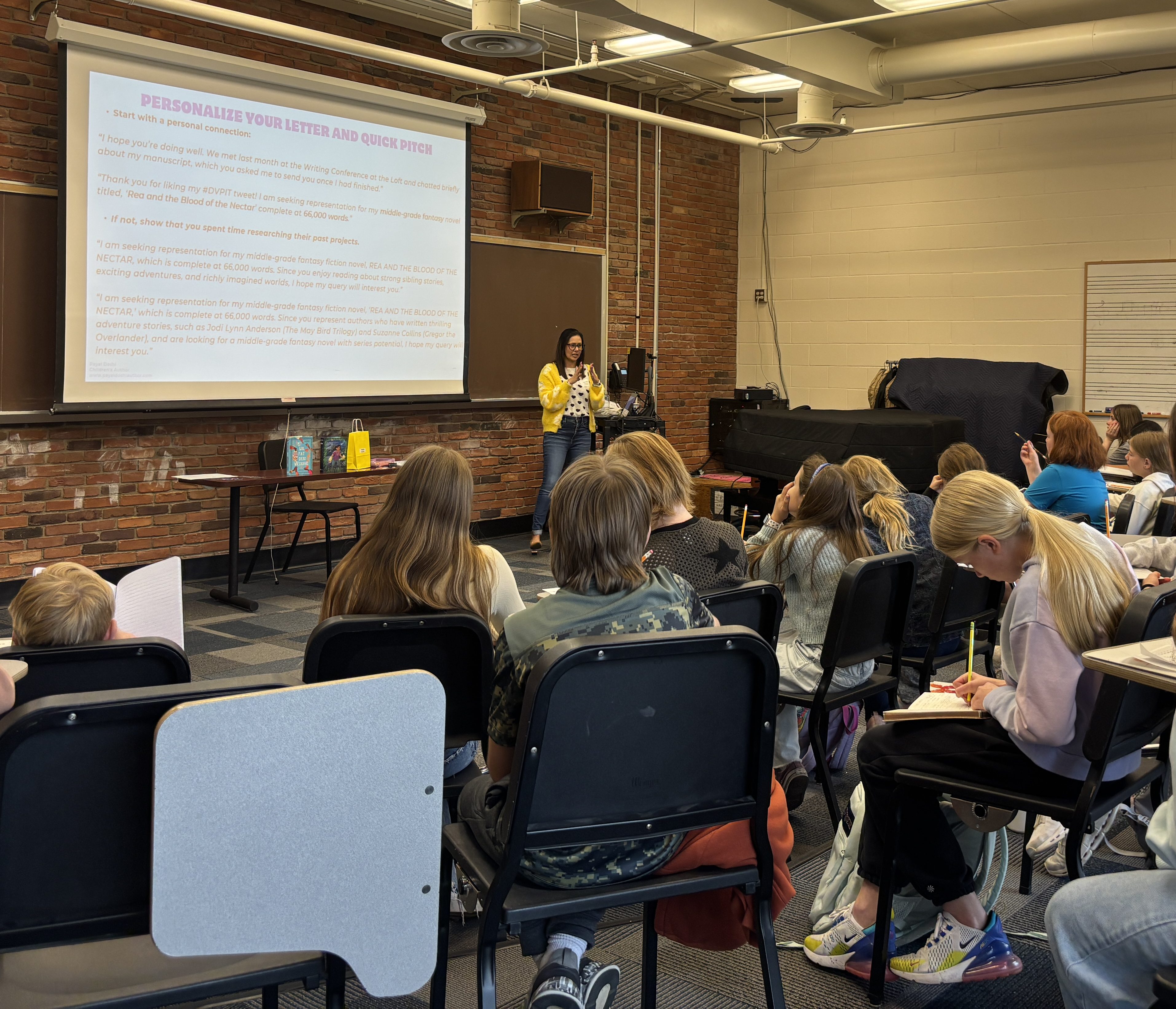 A classroom with students seated, listening to a presenter speaking at the front by a presentation screen.