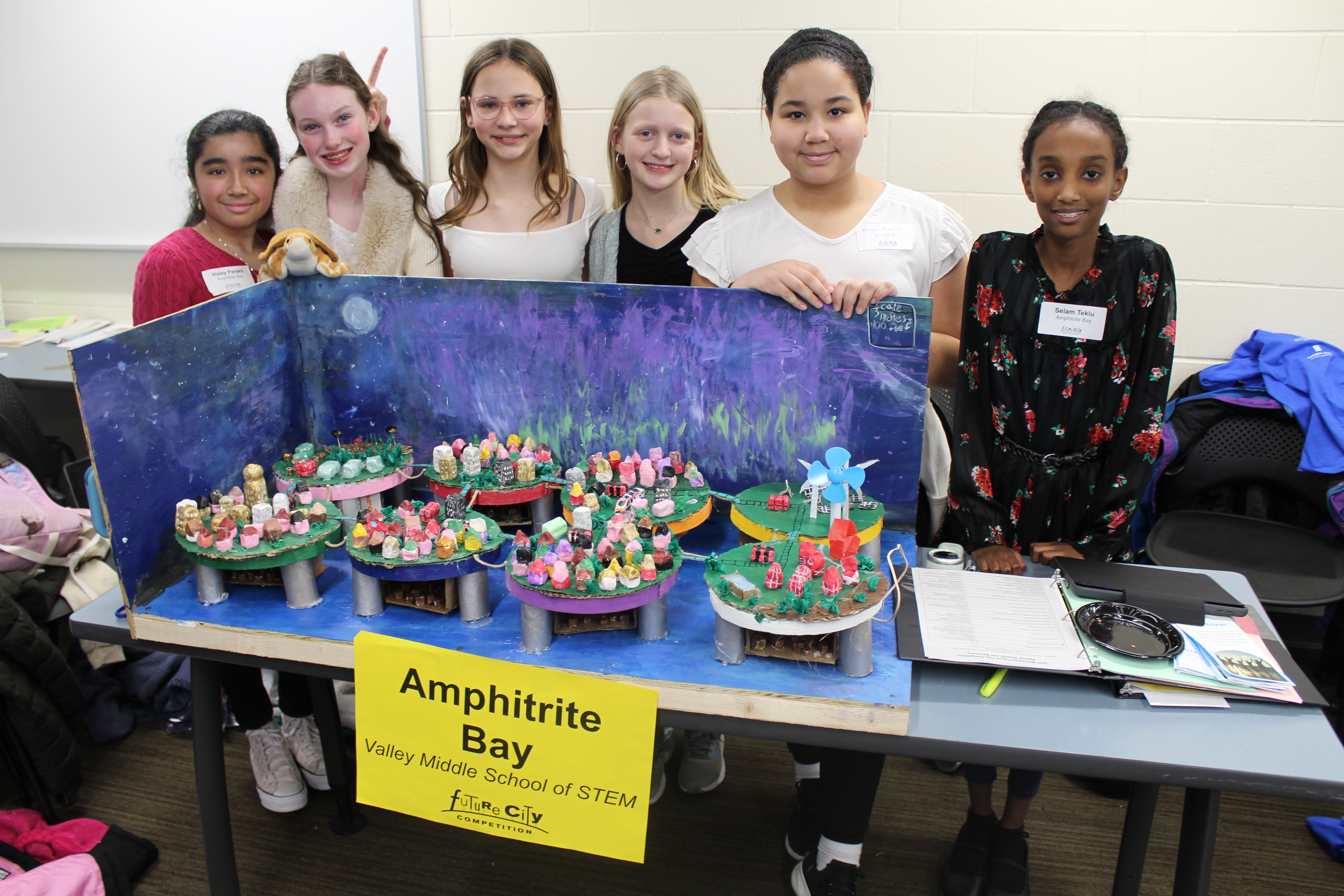 Six girls stand behind a colorful model called "Amphitrite Bay" at a school STEM event.