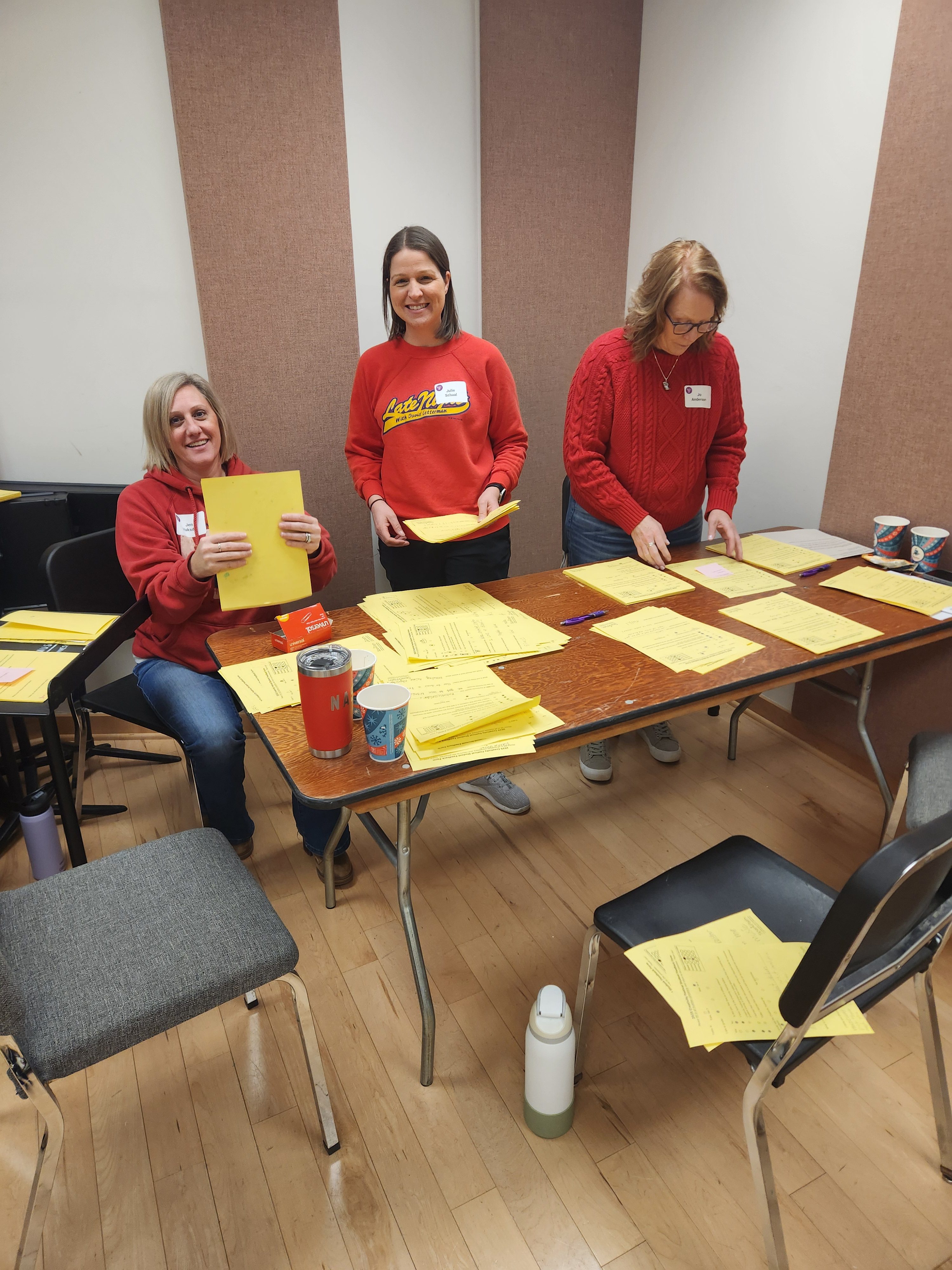 Three women sorting yellow papers at a table in a classroom, all wearing red sweaters and smiling.