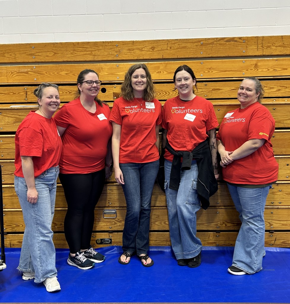 Five women in red volunteer shirts stand and smile in front of wooden bleachers in a gymnasium.