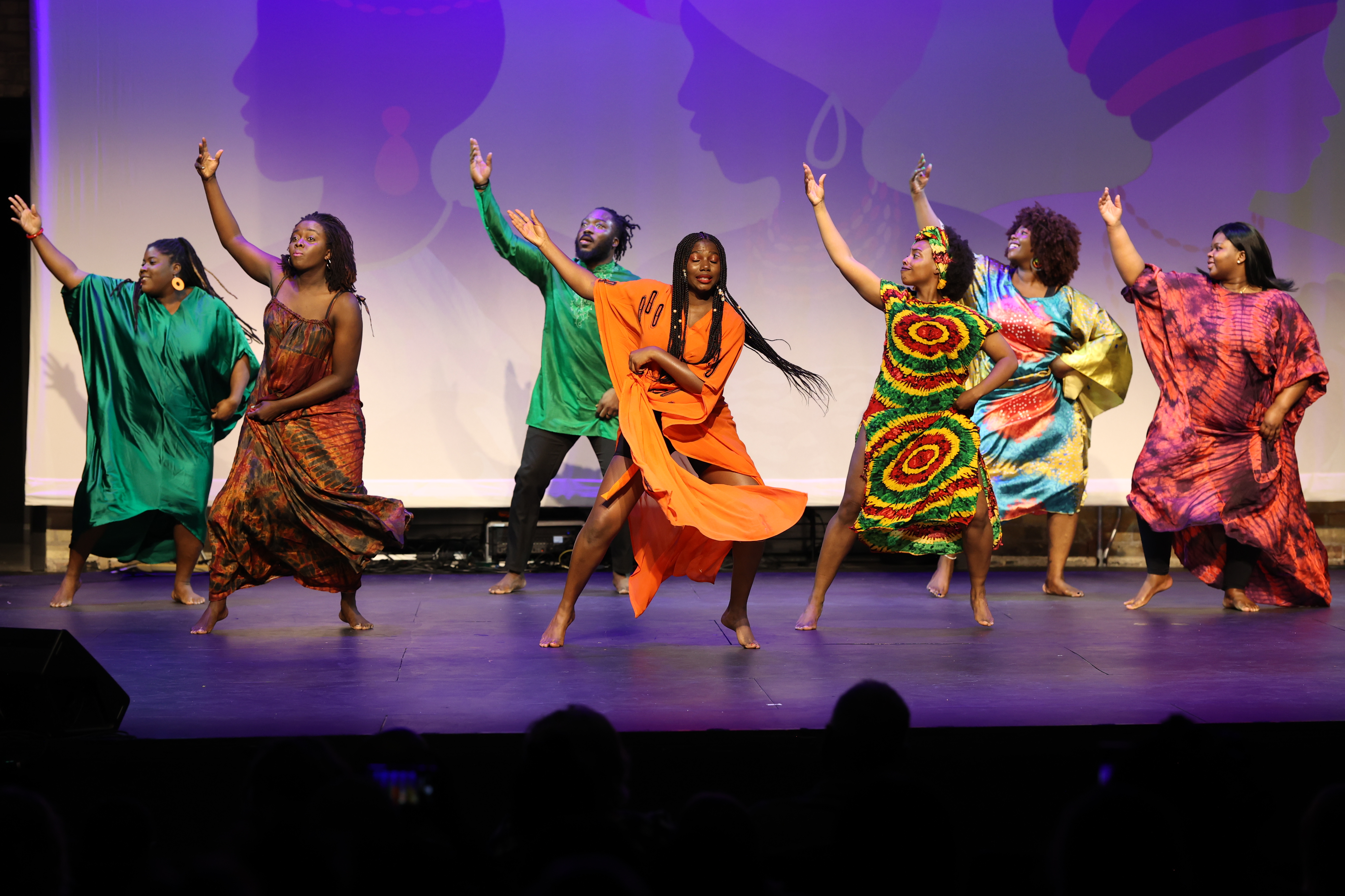 A group of dancers in colorful outfits perform on stage with arms raised.