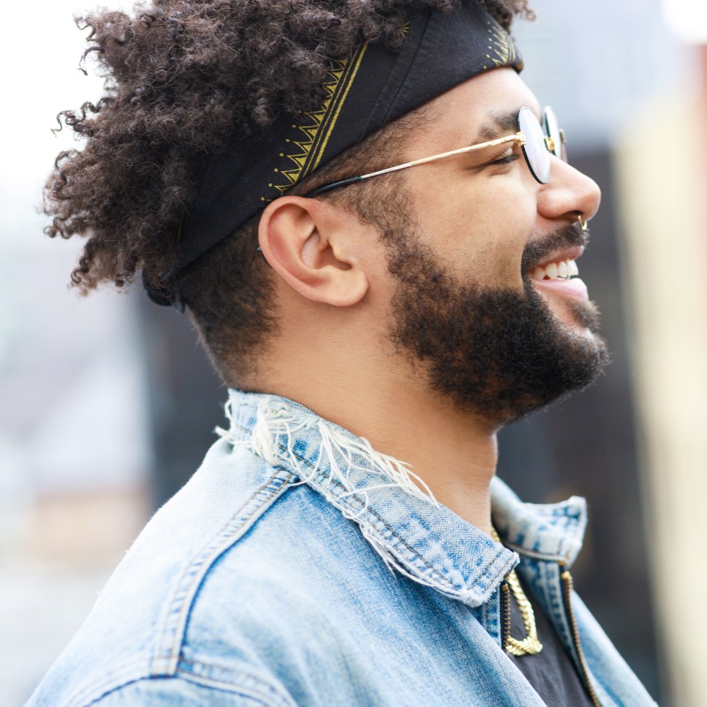 Smiling man in sunglasses and denim jacket, wearing a headband, in profile outdoors.