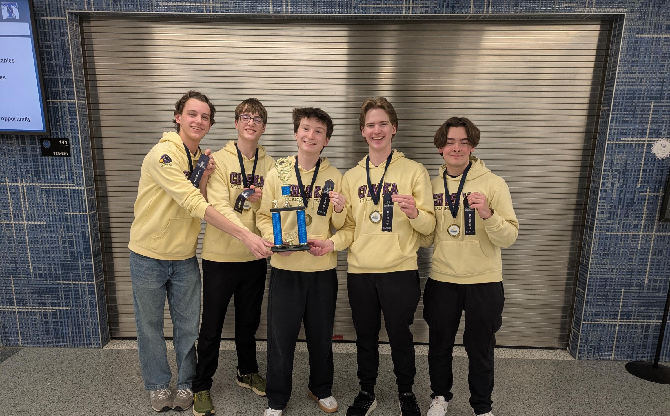Five students in yellow hoodies holding a trophy and medals, smiling in front of a metal shutter.