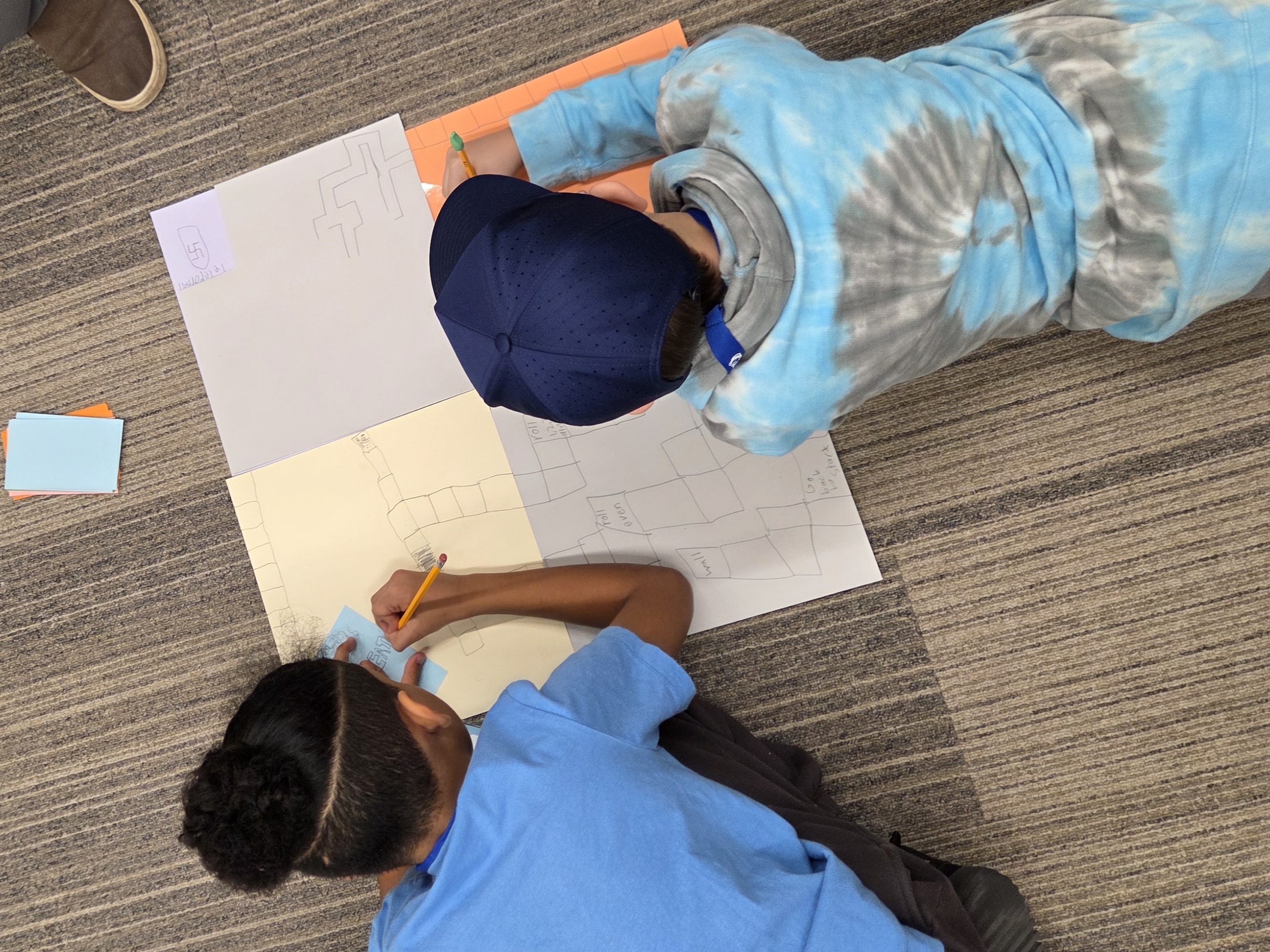 Two kids draw on a large sheet of paper on the floor, surrounded by colored paper, sticky notes, and scissors.