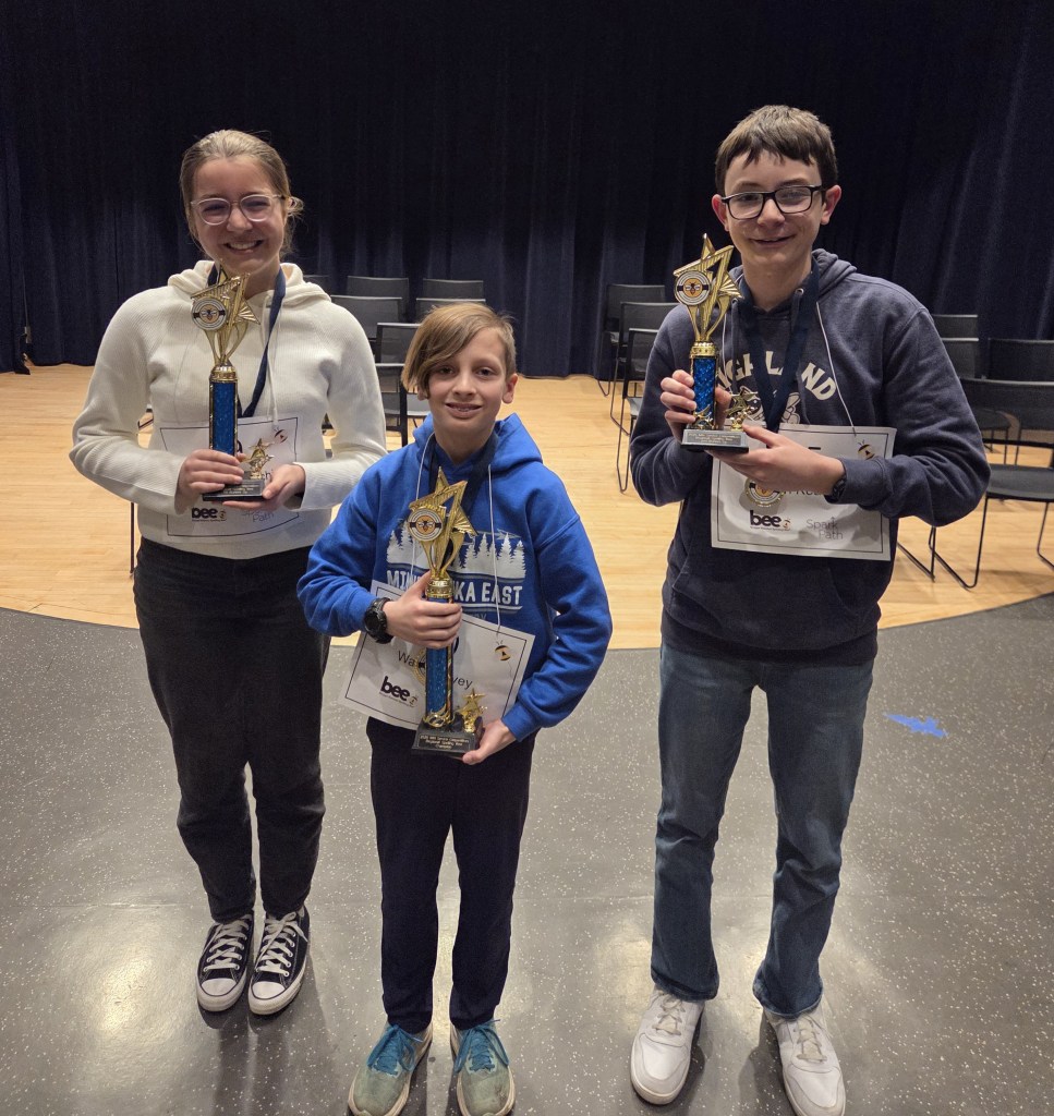 Three smiling kids holding trophies and certificates on a stage, wearing spelling bee contestant numbers.