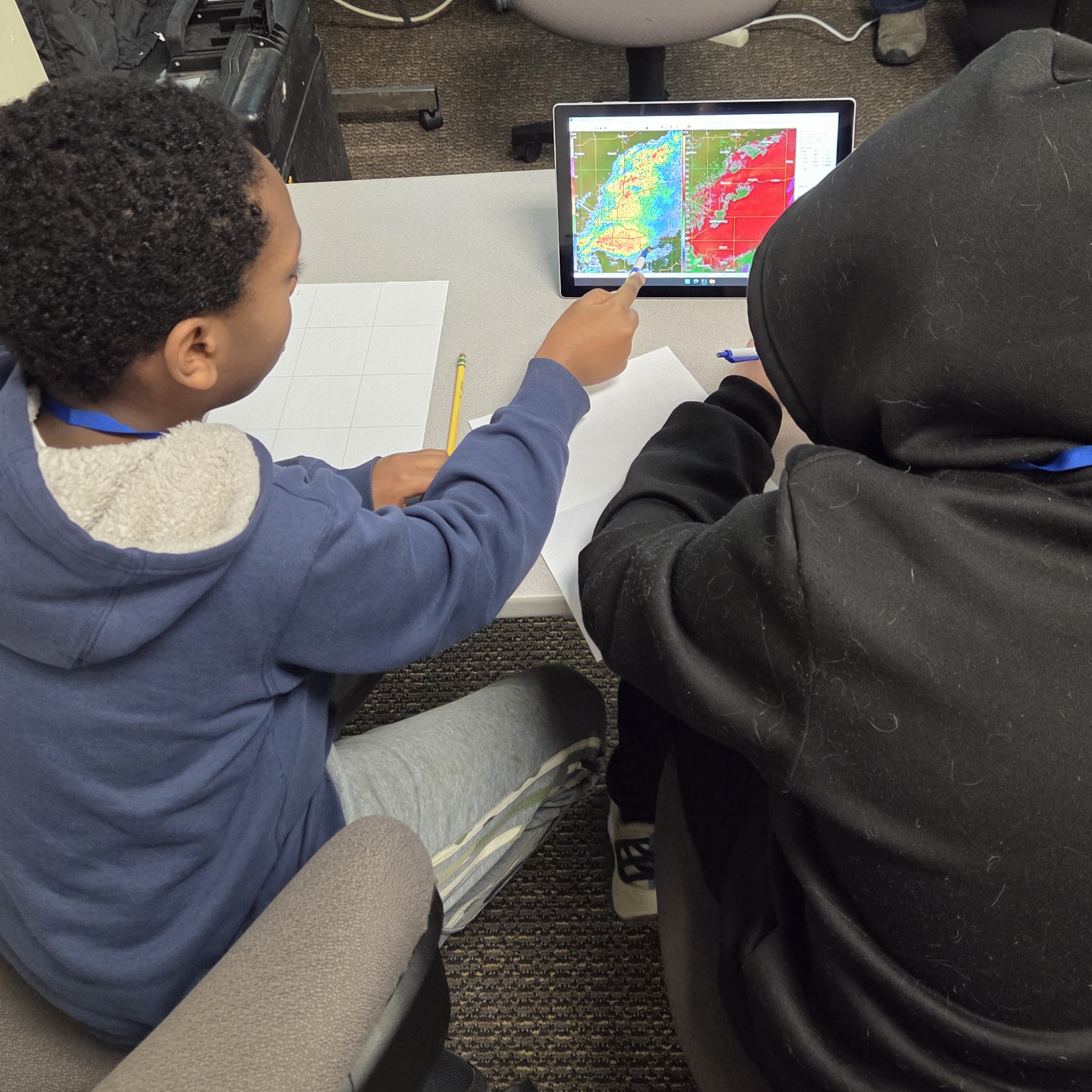 Two kids at a table analyze weather radar on a tablet, one pointing at the screen with a pencil.