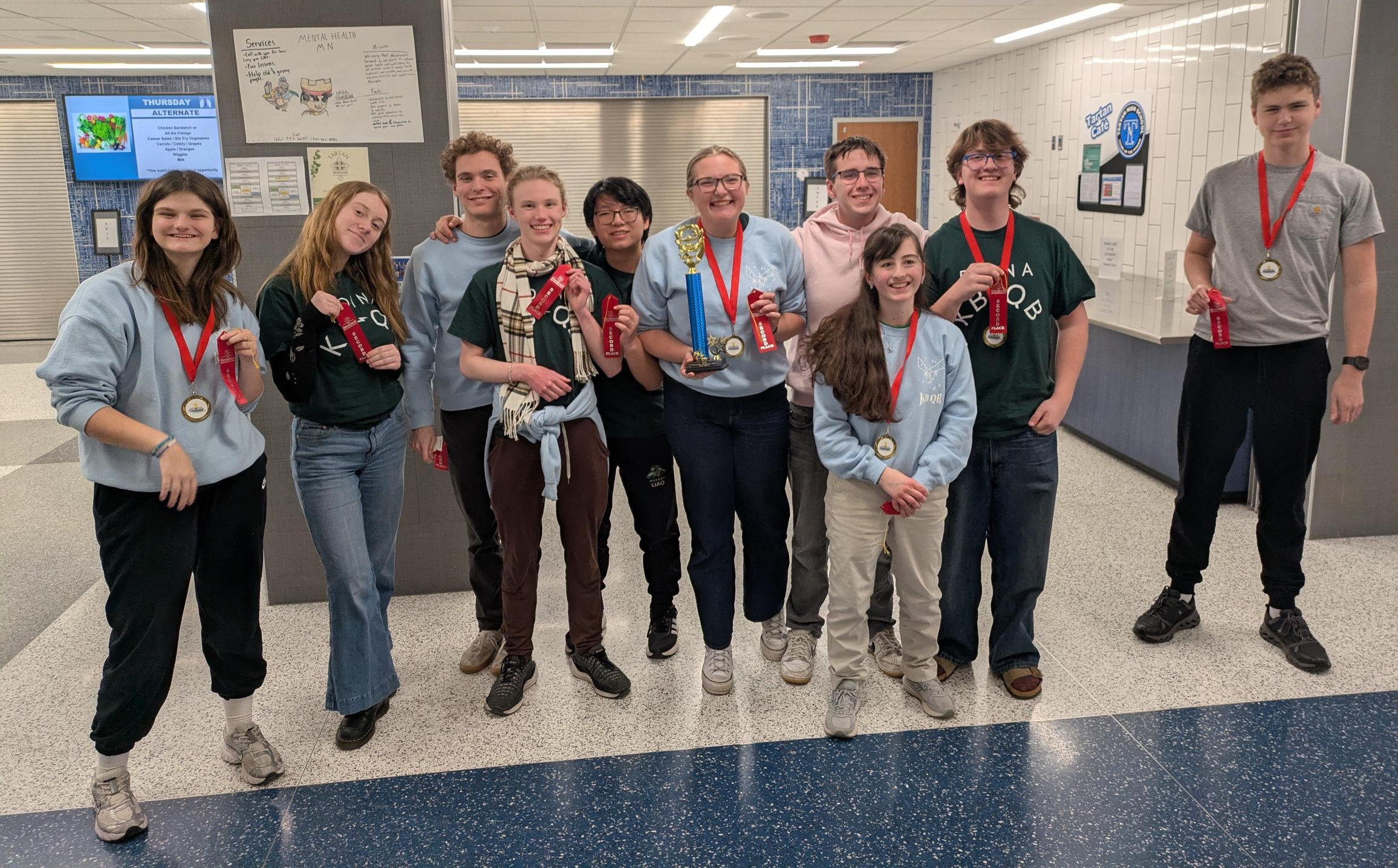 A group of smiling students wearing medals and holding red bottles pose together in a school hallway.