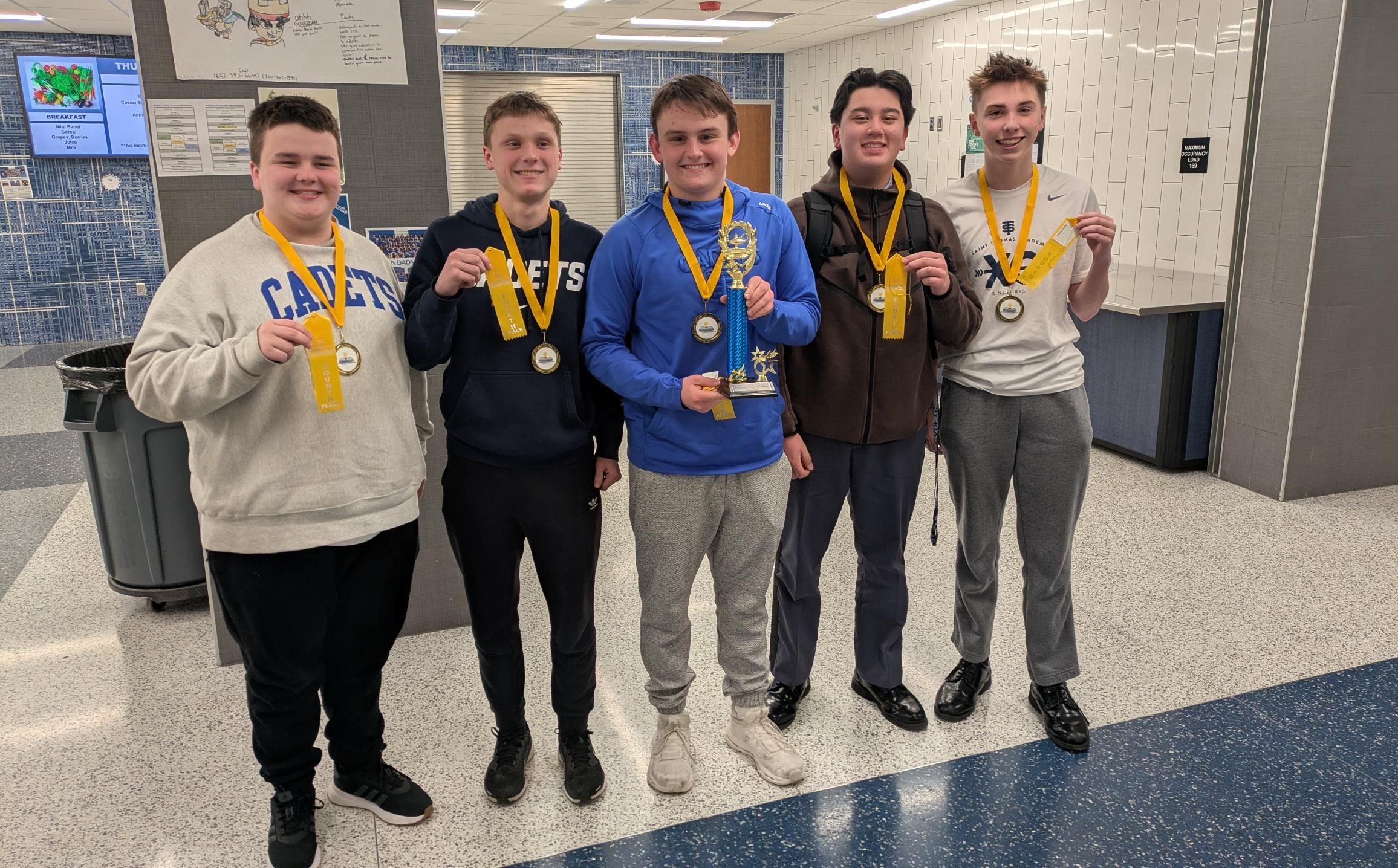 Five smiling students with medals pose indoors; one holds a trophy. They stand on a blue and white tiled floor.