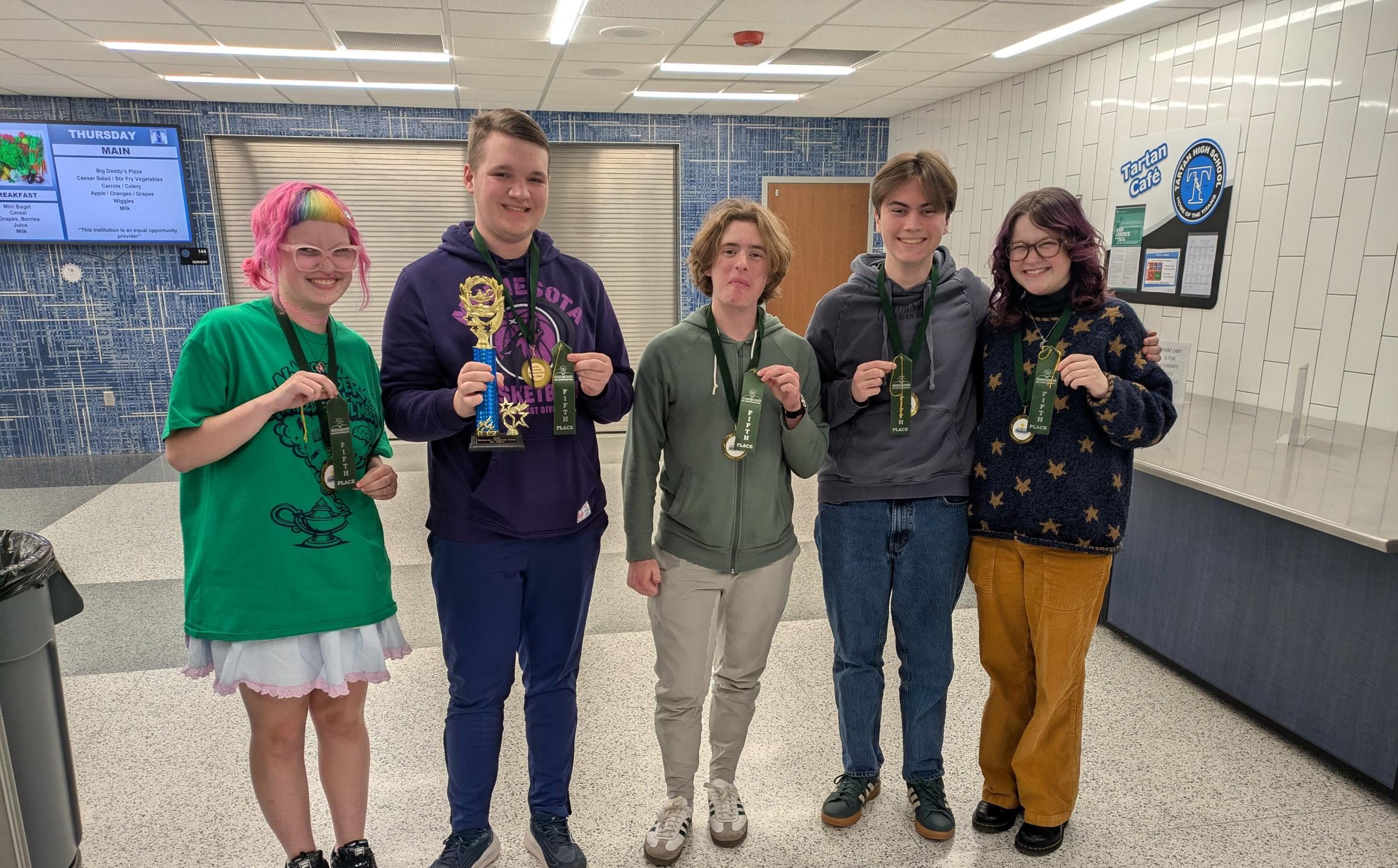 Five smiling teens stand indoors holding medals and a trophy, celebrating their achievements together.