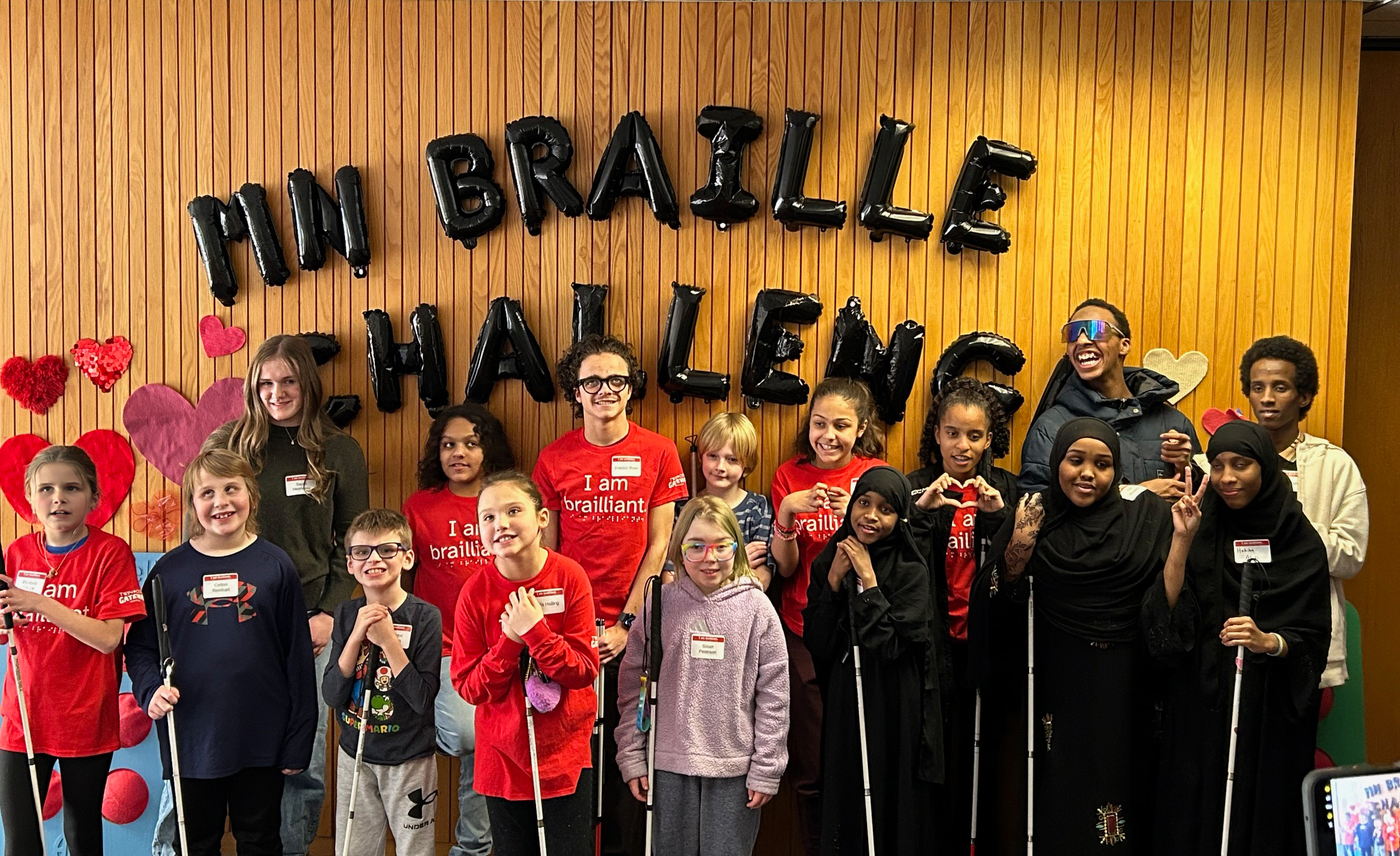 A group of kids and adults pose, some with canes, in front of "MN Braille Challenge" balloon letters.