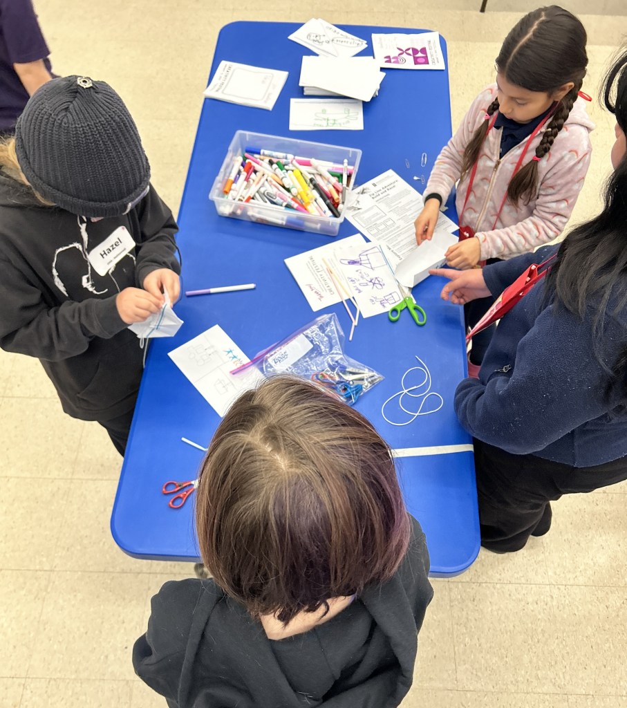 Four children standing around a blue table working on arts and crafts with papers, markers, and scissors.