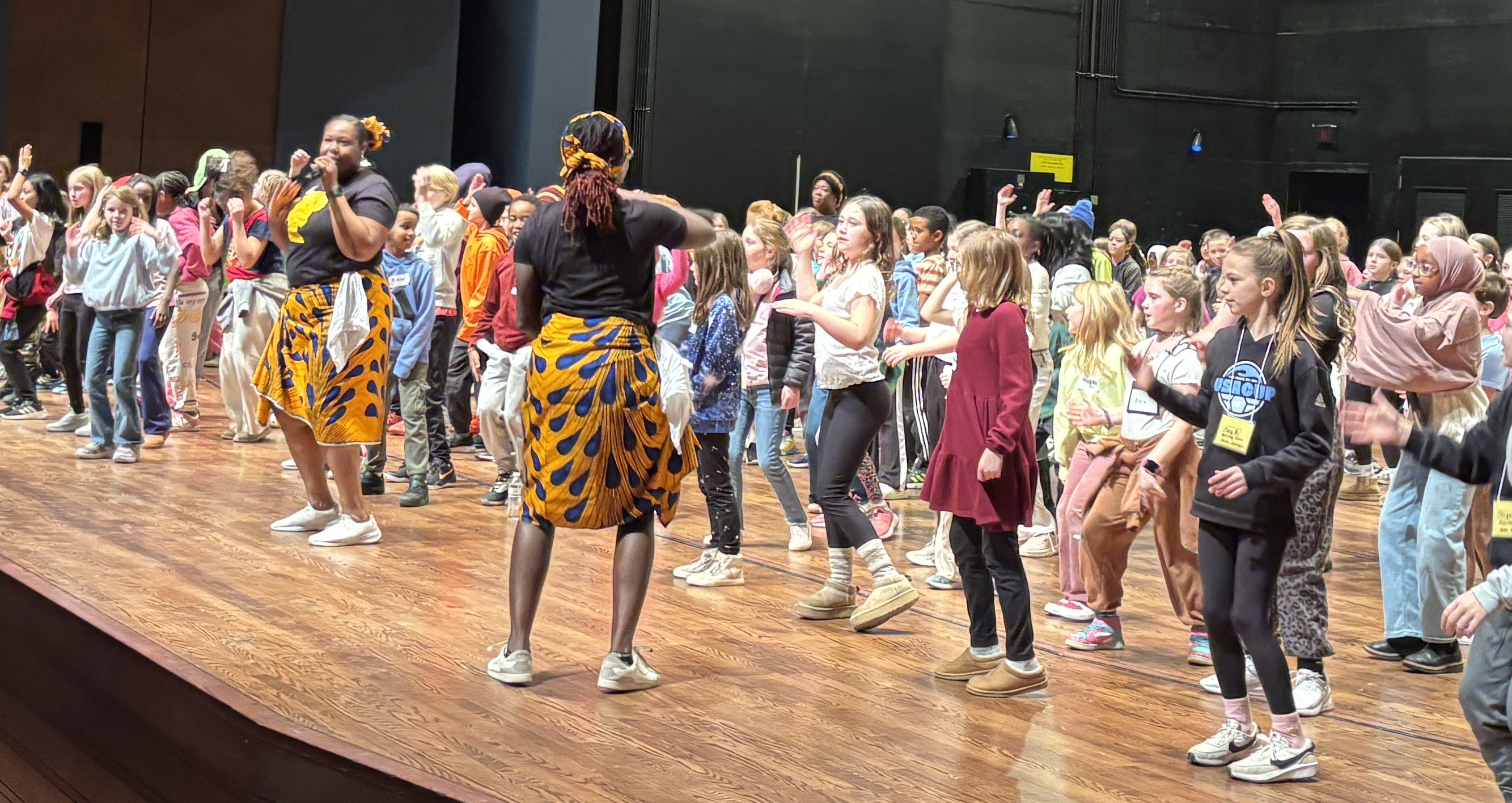 Two women lead a large group of children in a lively dance on a stage with a wooden floor.