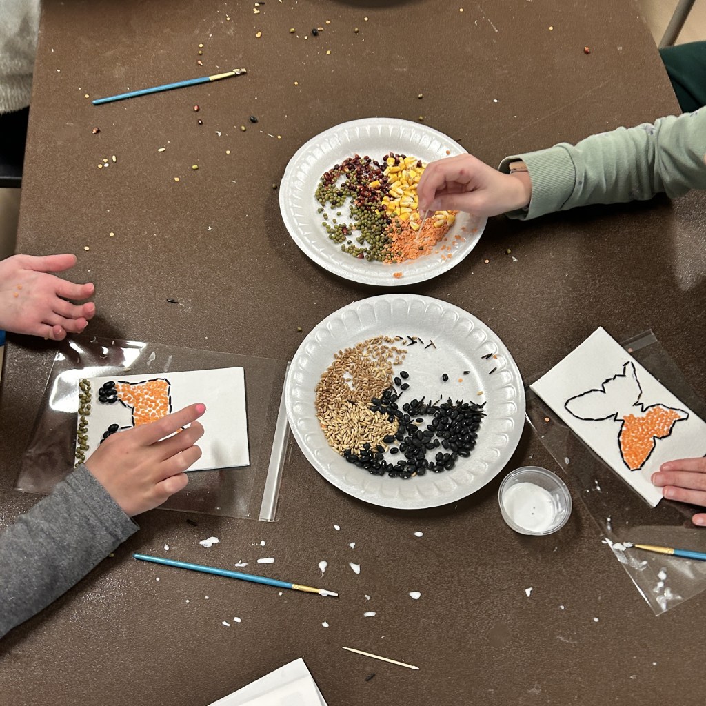 Three children use seeds to make crop art at a brown table with plates of supplies.