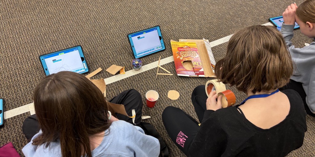 Three kids sitting on the floor with tablets, cardboard, tape, and STEM materials, working on a project.
