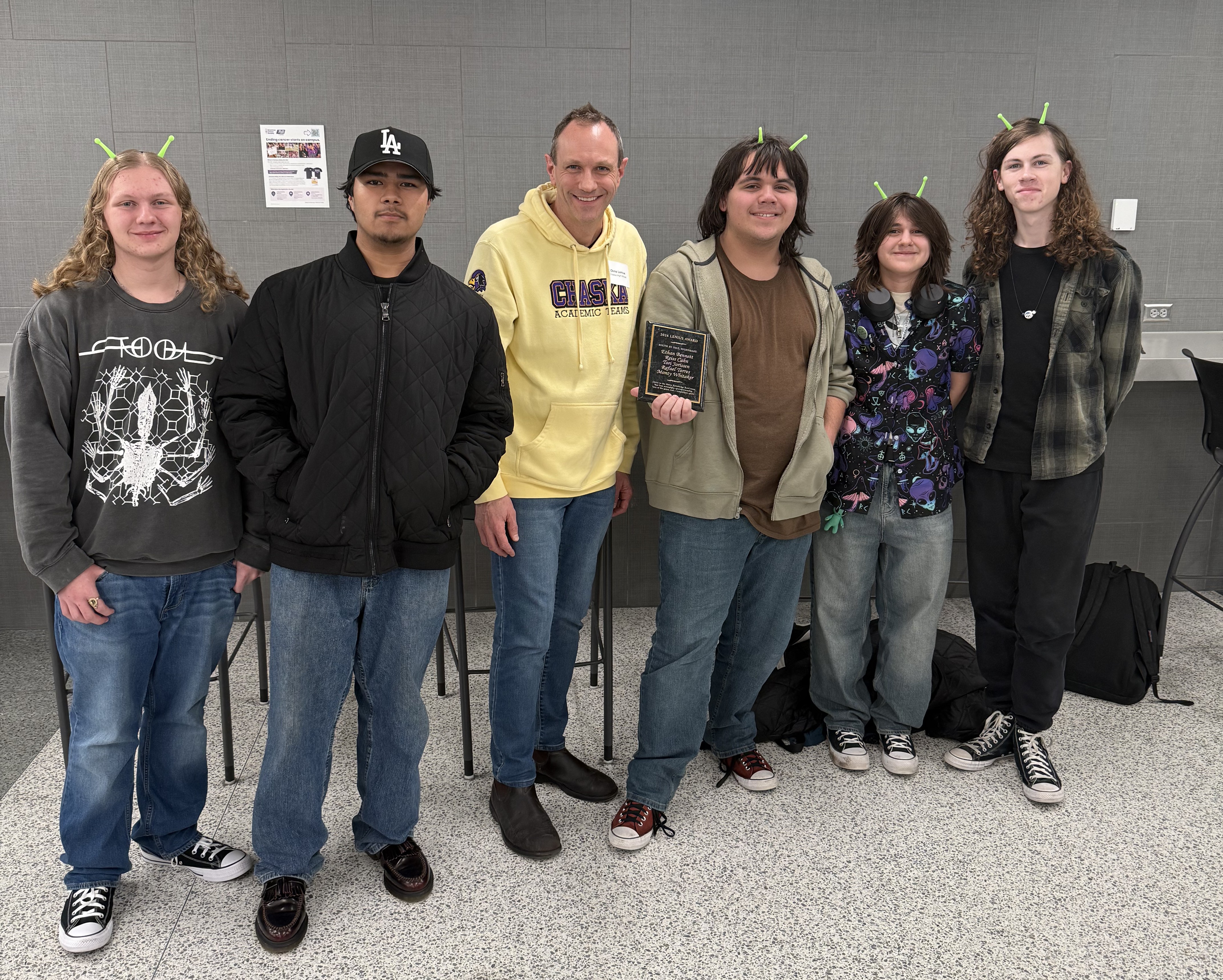 Six people stand together indoors, five wearing Shrek ears and one holding a plaque, all smiling at the camera.