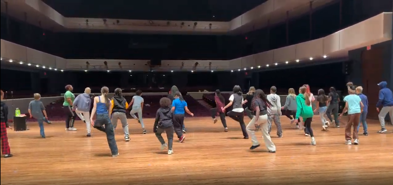 A group of people practice a dance routine on a large theater stage facing an empty auditorium.