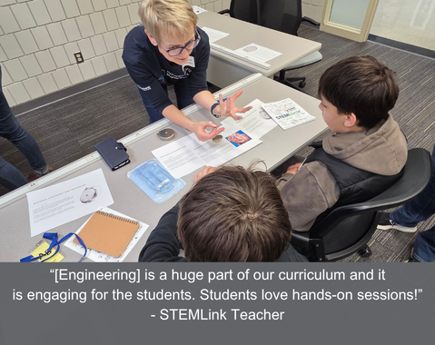 A teacher demonstrates a science activity to two students at a table, with STEM materials and papers around.