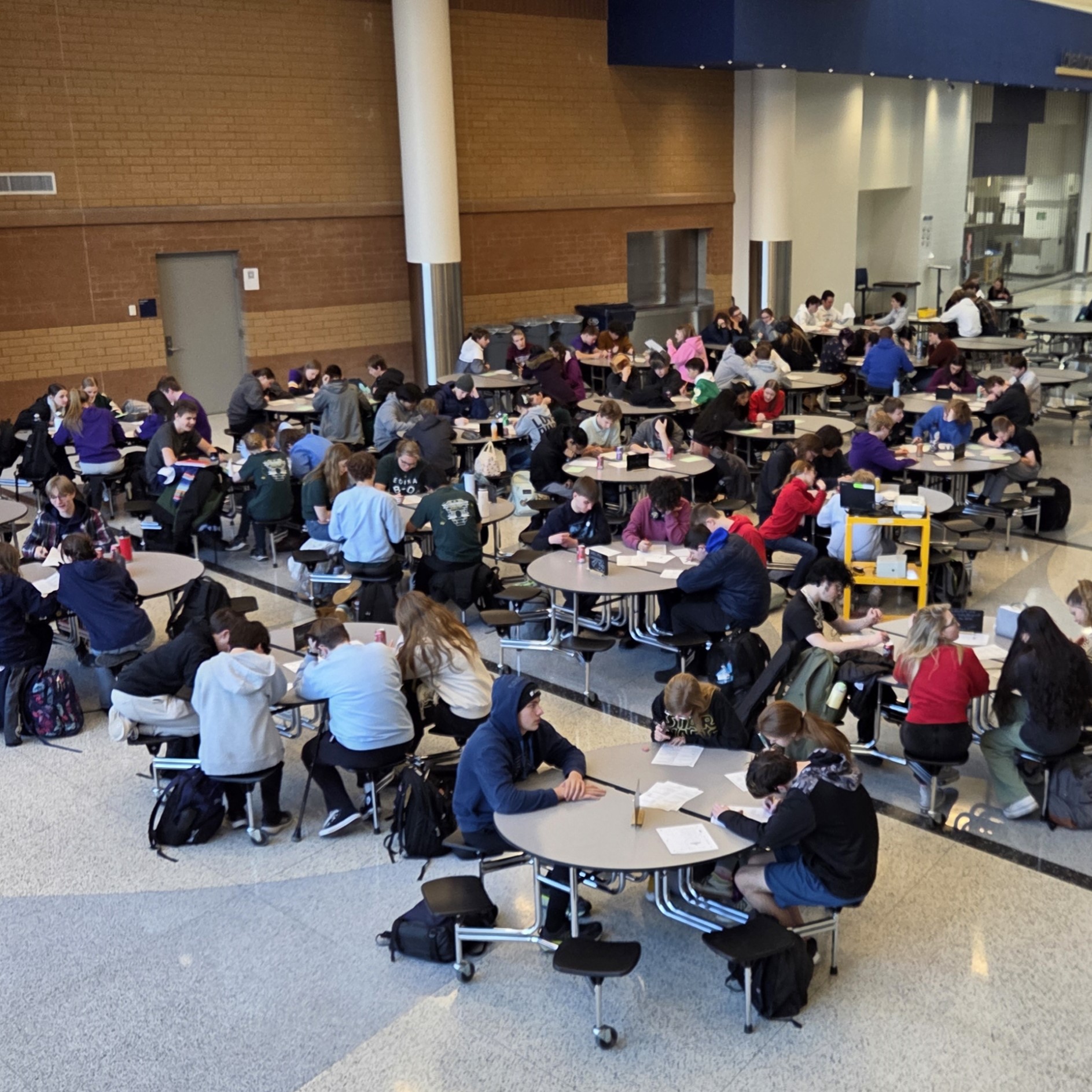Large group of students sitting at round tables in a cafeteria, working on the written round at Knowledge Bowl