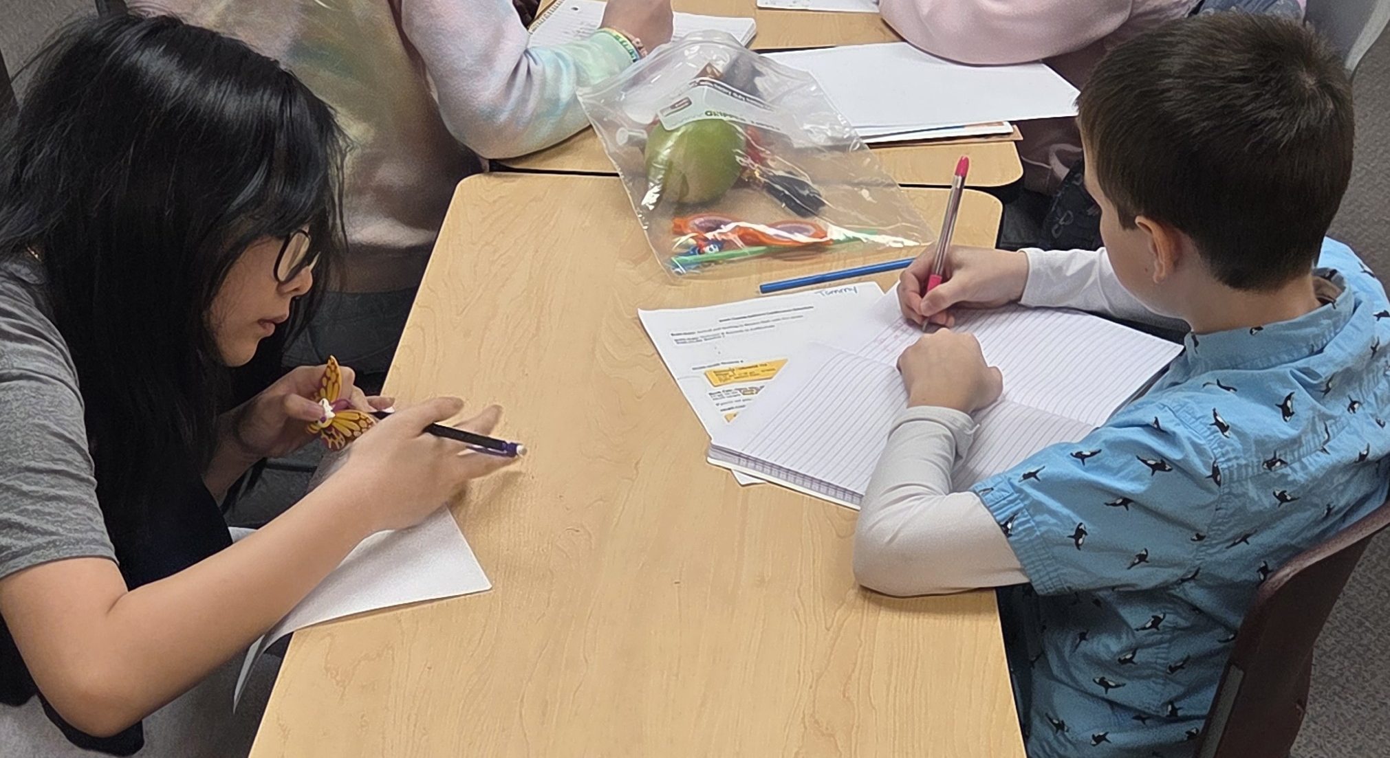 Four students sit at tables, writing or drawing on paper with pencils and markers at the young authors conference