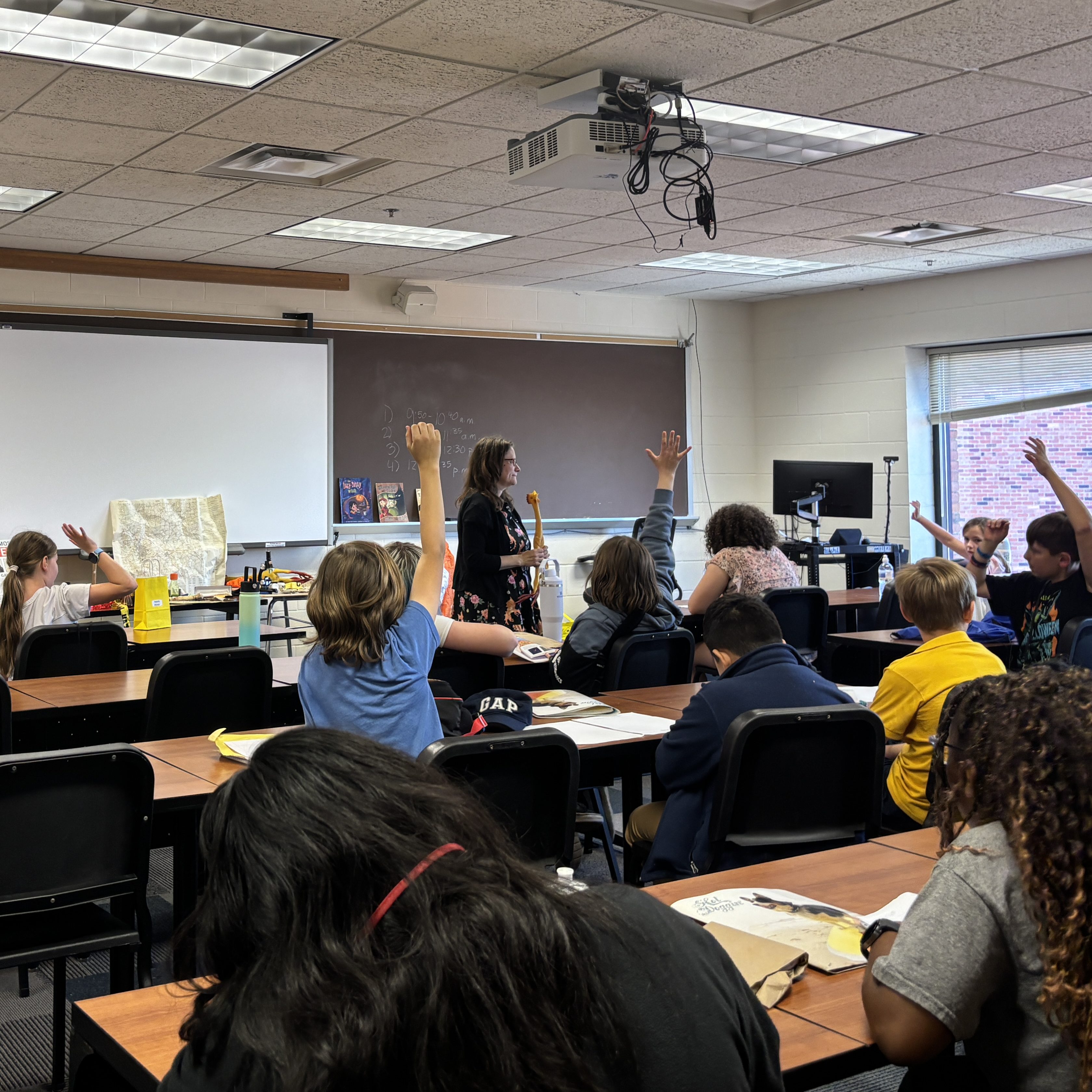 Students in a session raise their hands while an author stands at the front near a whiteboard.
