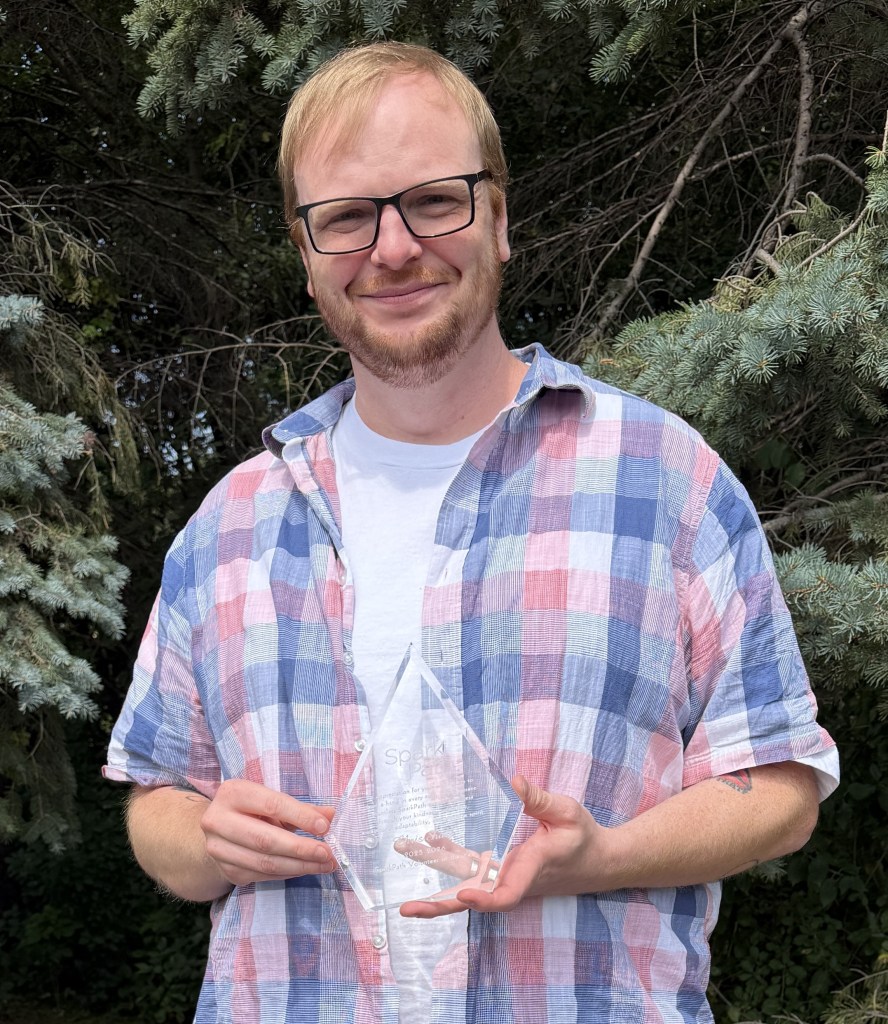A person with glasses holds a clear triangular award, standing outdoors in front of pine trees.