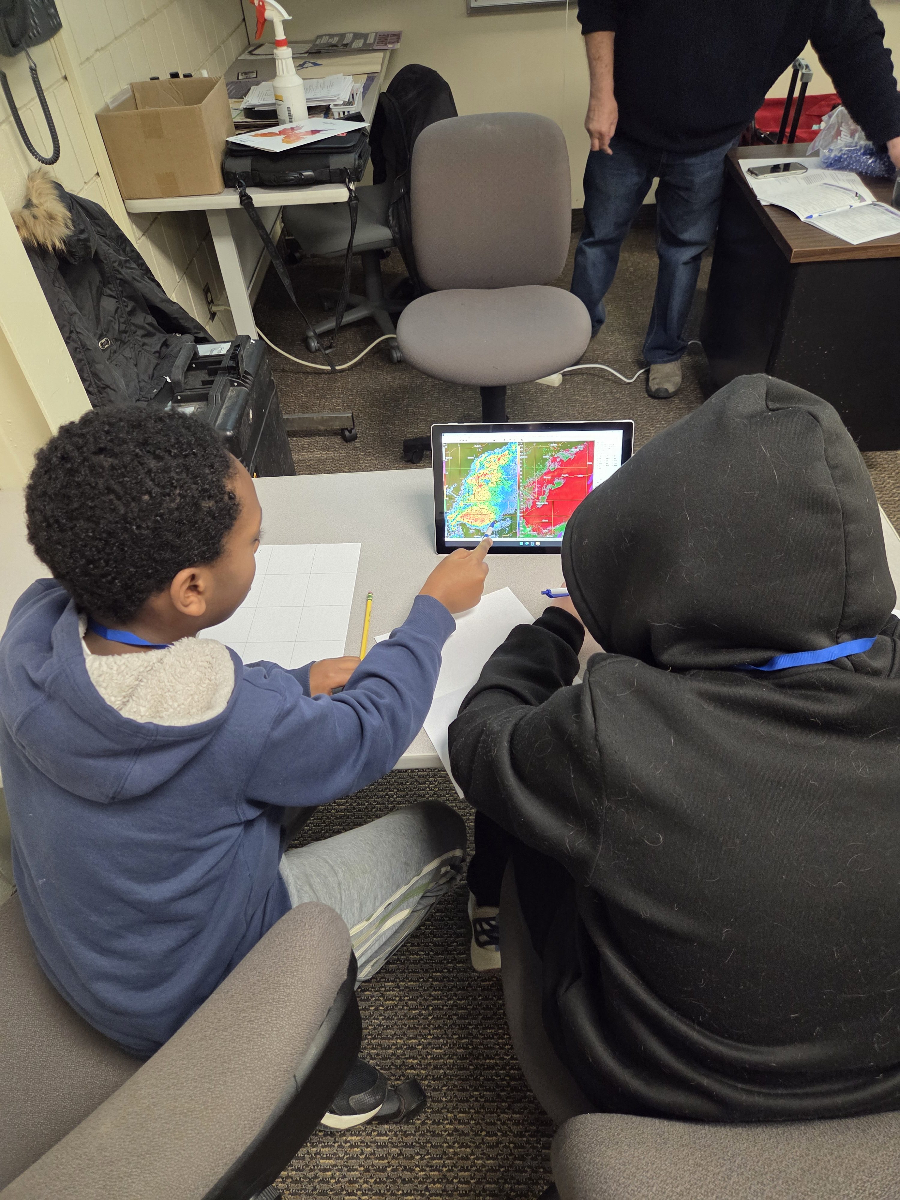Two children look at a colorful weather map on a tablet, discussing and pointing at the screen together.