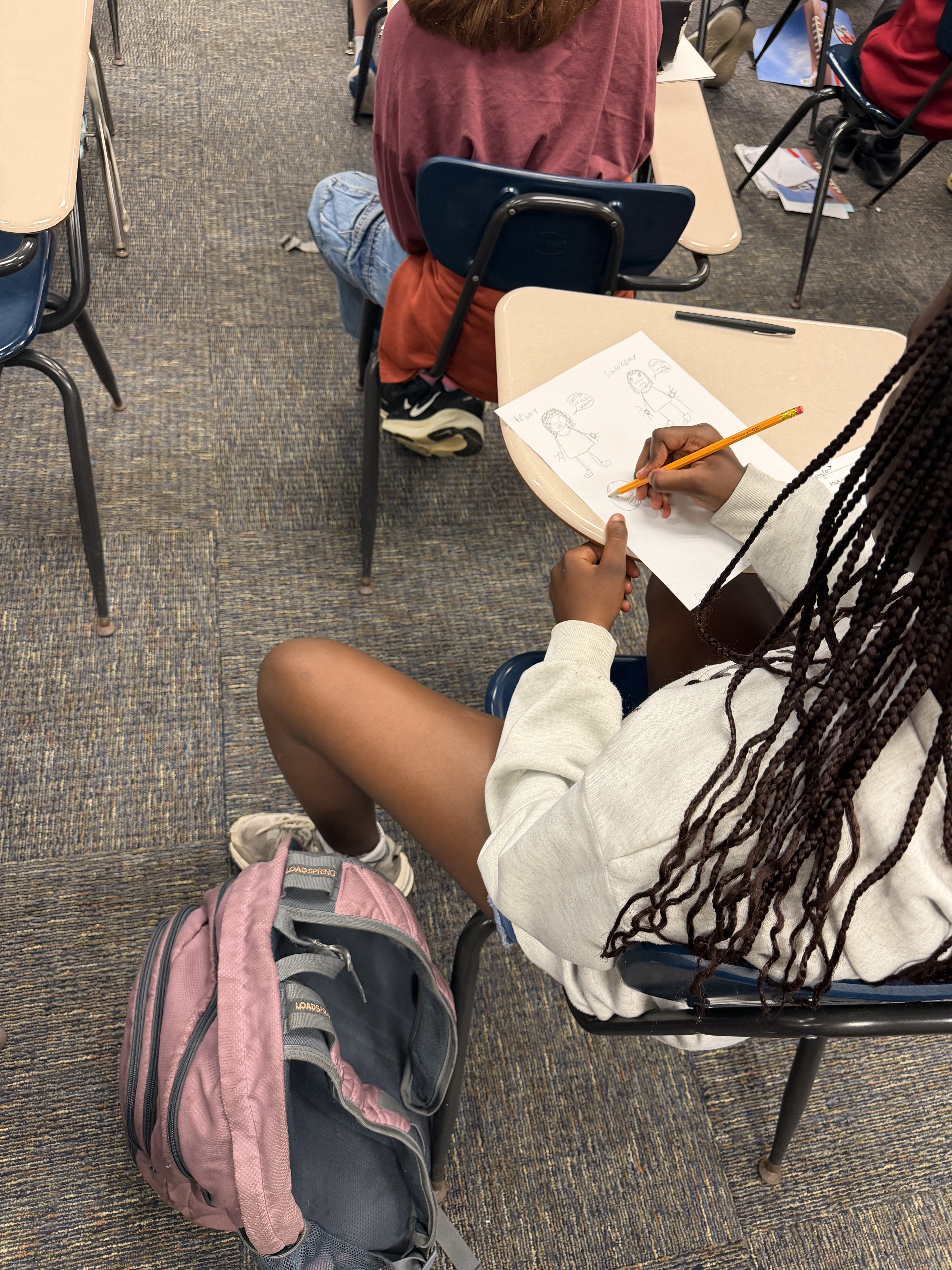 Student drawing on paper at a desk with a backpack on the floor in a classroom setting.
